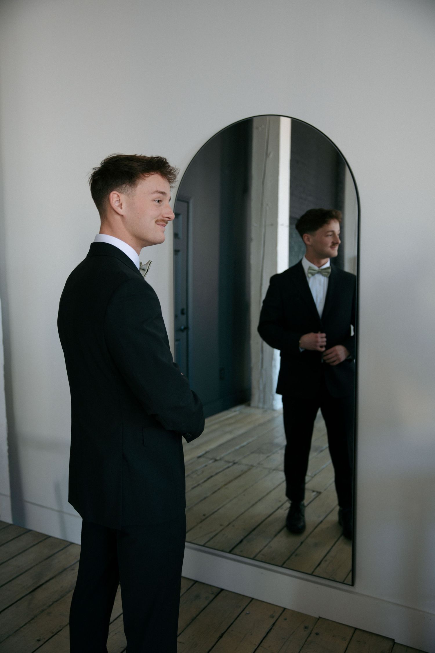 Young groom in black tuxedo admiring himself in an arched full-length mirror before his wedding ceremony.