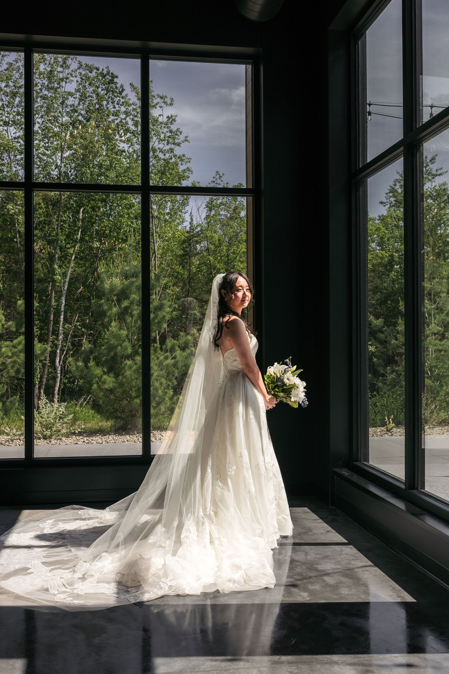 Bride in a flowing lace gown and long veil holds a bouquet while standing by tall windows in natural light.