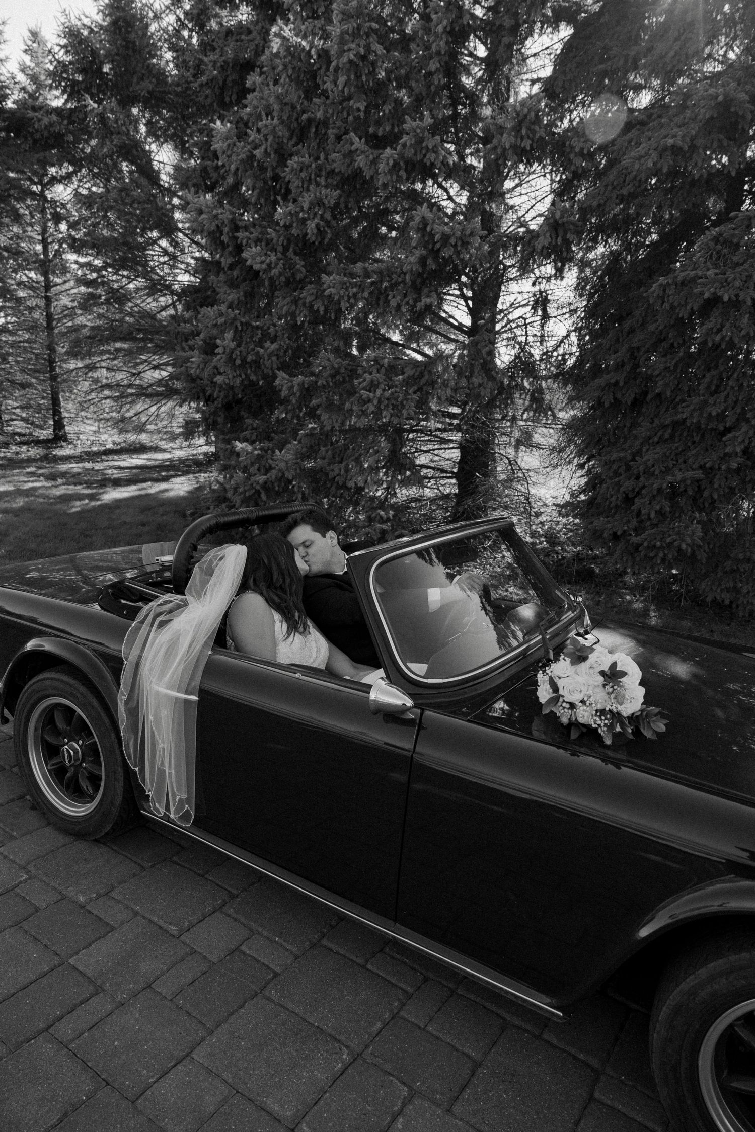 Black and white photo of newlyweds sharing a kiss in a vintage convertible car surrounded by trees.