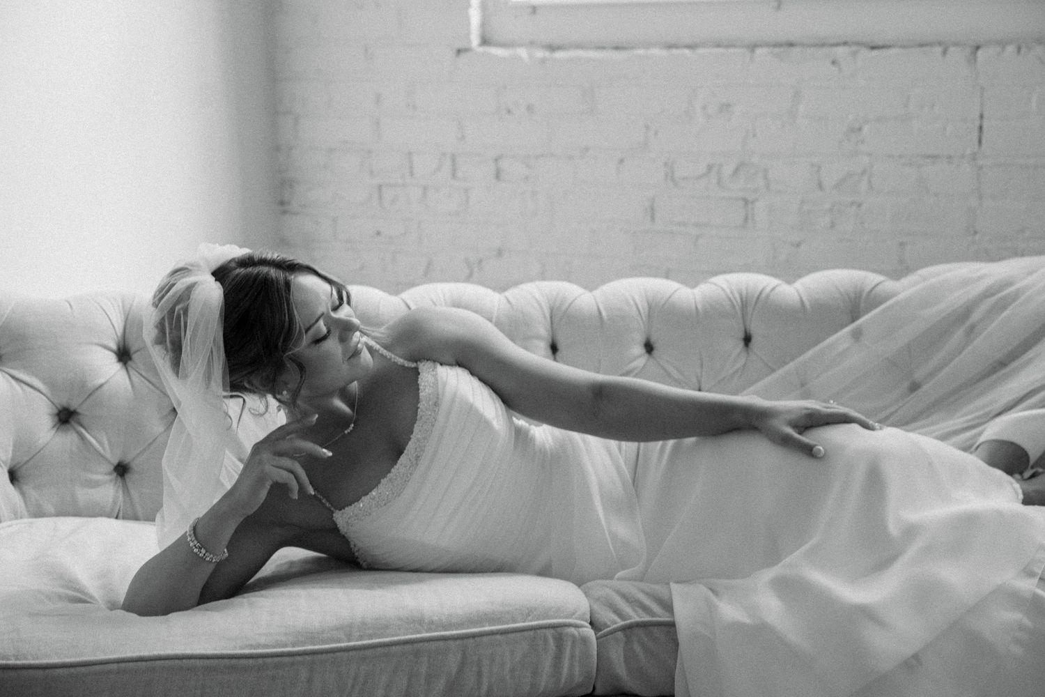 Bride in white gown and veil reclining elegantly on a tufted sofa in a black and white photo.