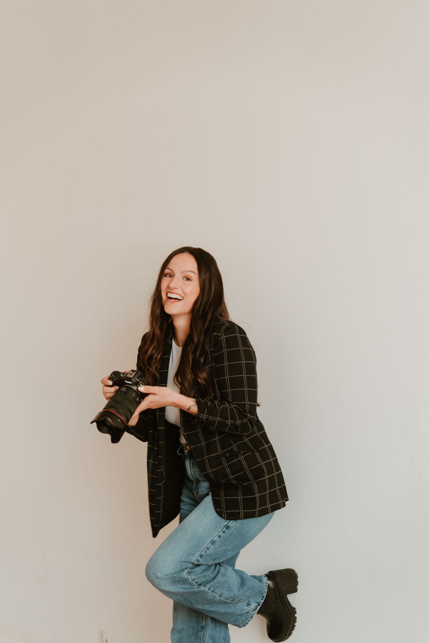 Smiling woman holding a DSLR camera, wearing a plaid blazer and jeans, posing playfully against a light gray wall.
