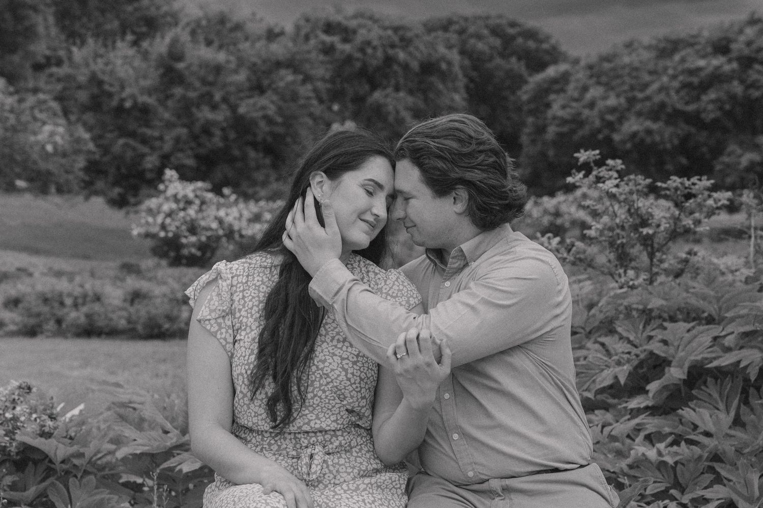 Black and white photo of a romantic couple sharing an intimate moment outdoors in a lush garden setting.