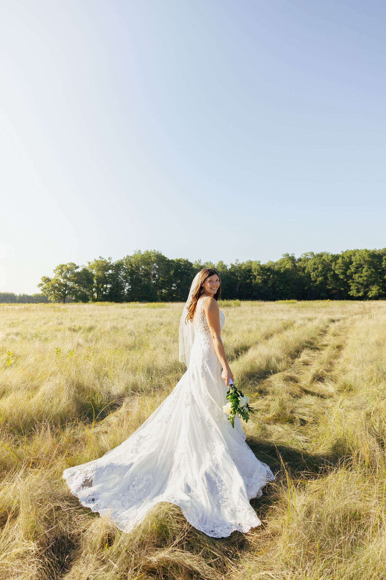 Elegant bride in lace gown with train holding bouquet in golden sunlit field.