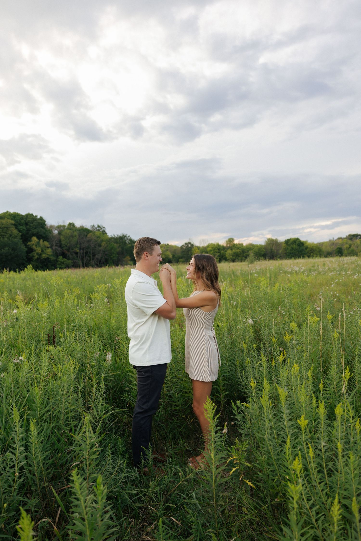Couple holding hands and gazing at each other in a lush green meadow under cloudy skies.