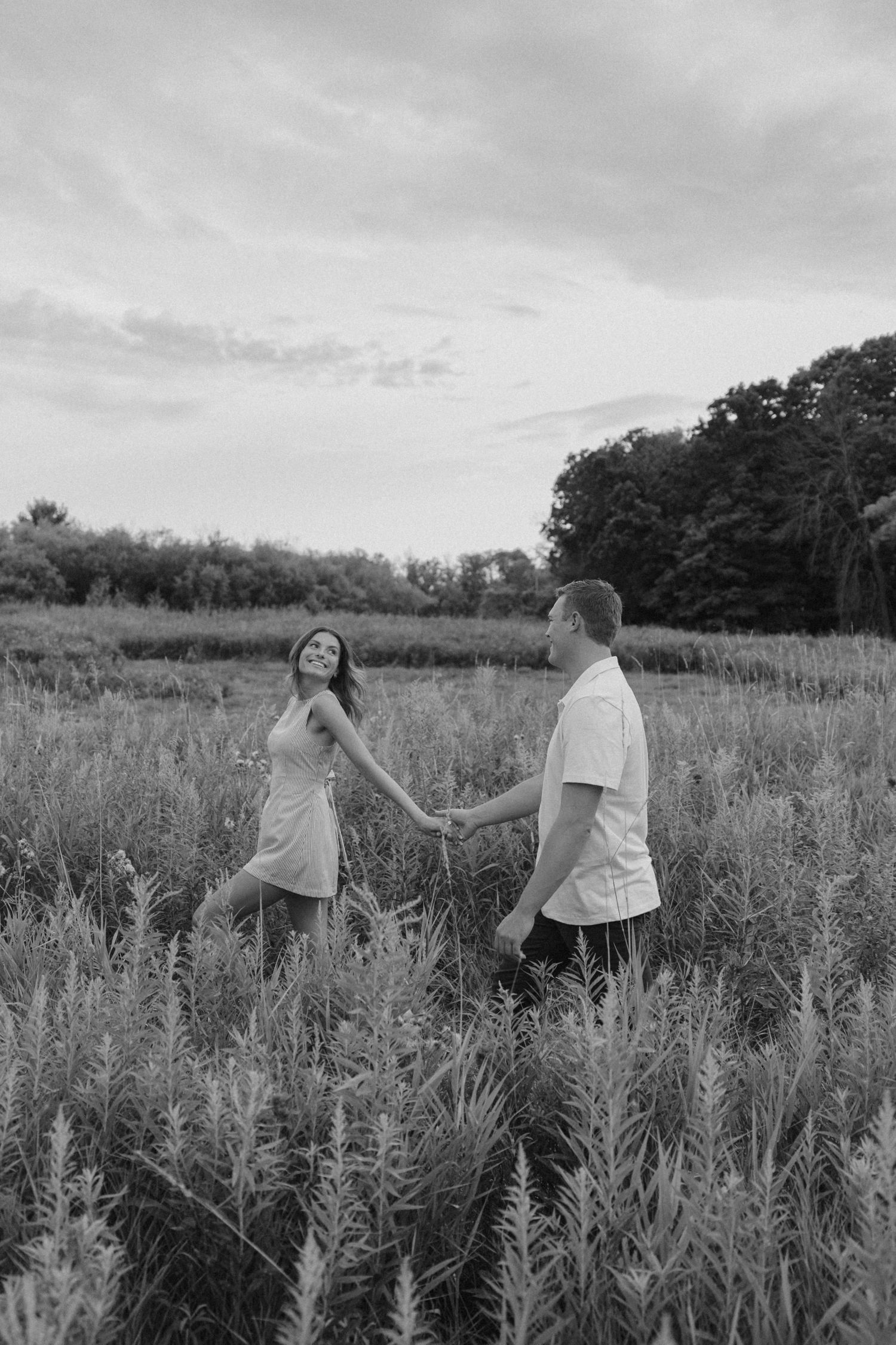 Black and white photo of a couple holding hands and walking through a lush green meadow.