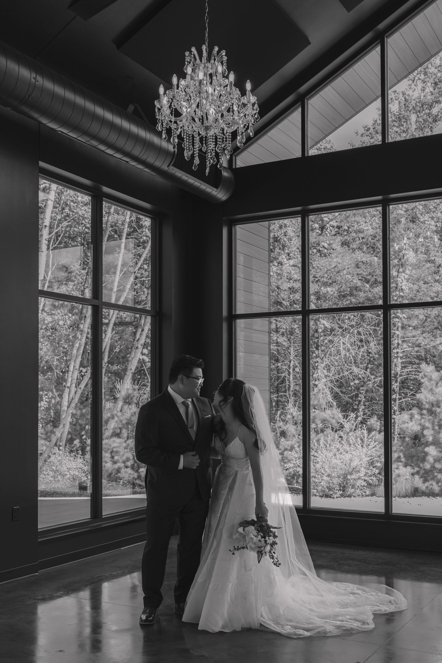 Black and white photo of bride and groom standing together under crystal chandelier with large windows in elegant venue.