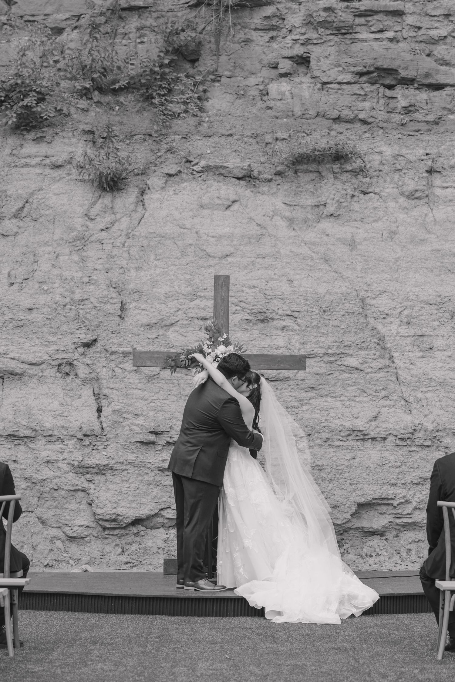 Bride and groom share first kiss at outdoor wedding ceremony with rustic stone wall and wooden cross backdrop.