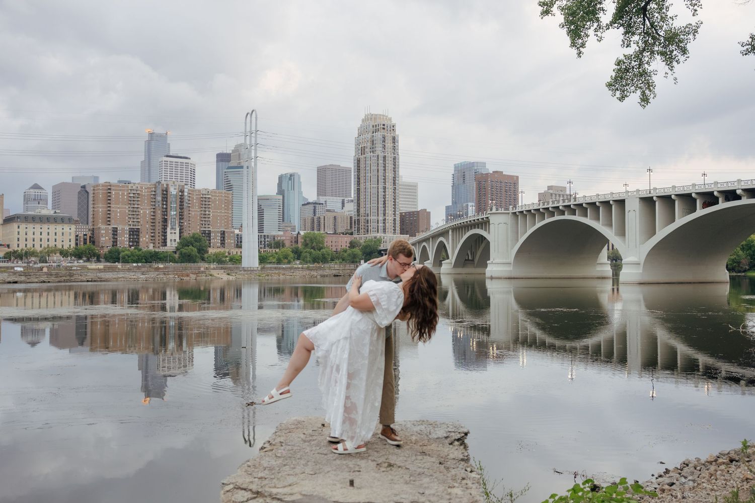 Couple sharing a romantic dip pose by the Mississippi River with Minneapolis skyline and bridge reflection in background.