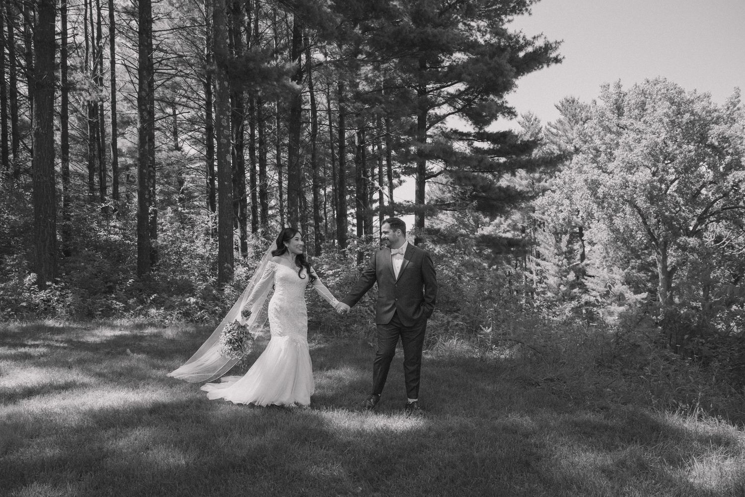 Black and white photo of bride and groom holding hands walking through sunlit forest.