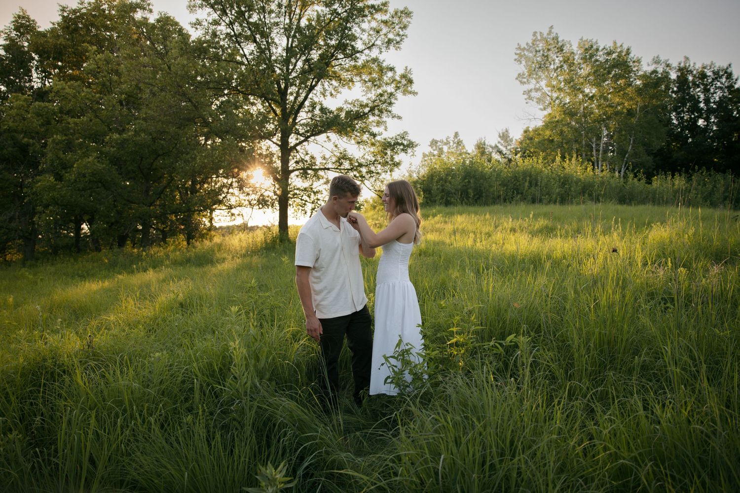 Couple standing in lush green field at golden hour, woman in white dress facing man in white shirt near trees.