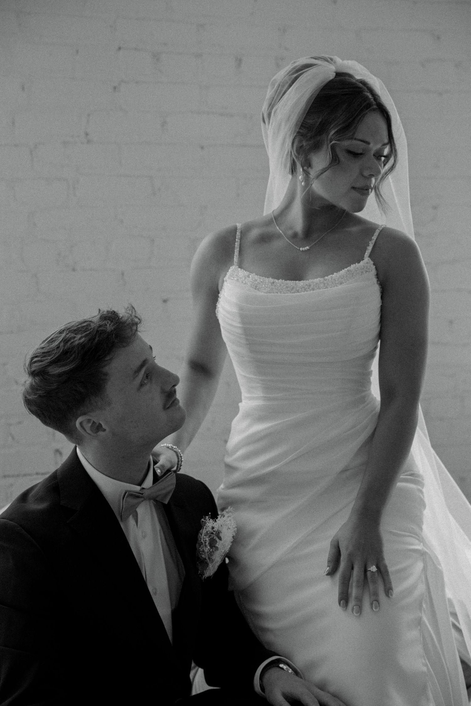 Black and white photo of groom kneeling before bride in elegant wedding gown and veil indoors.