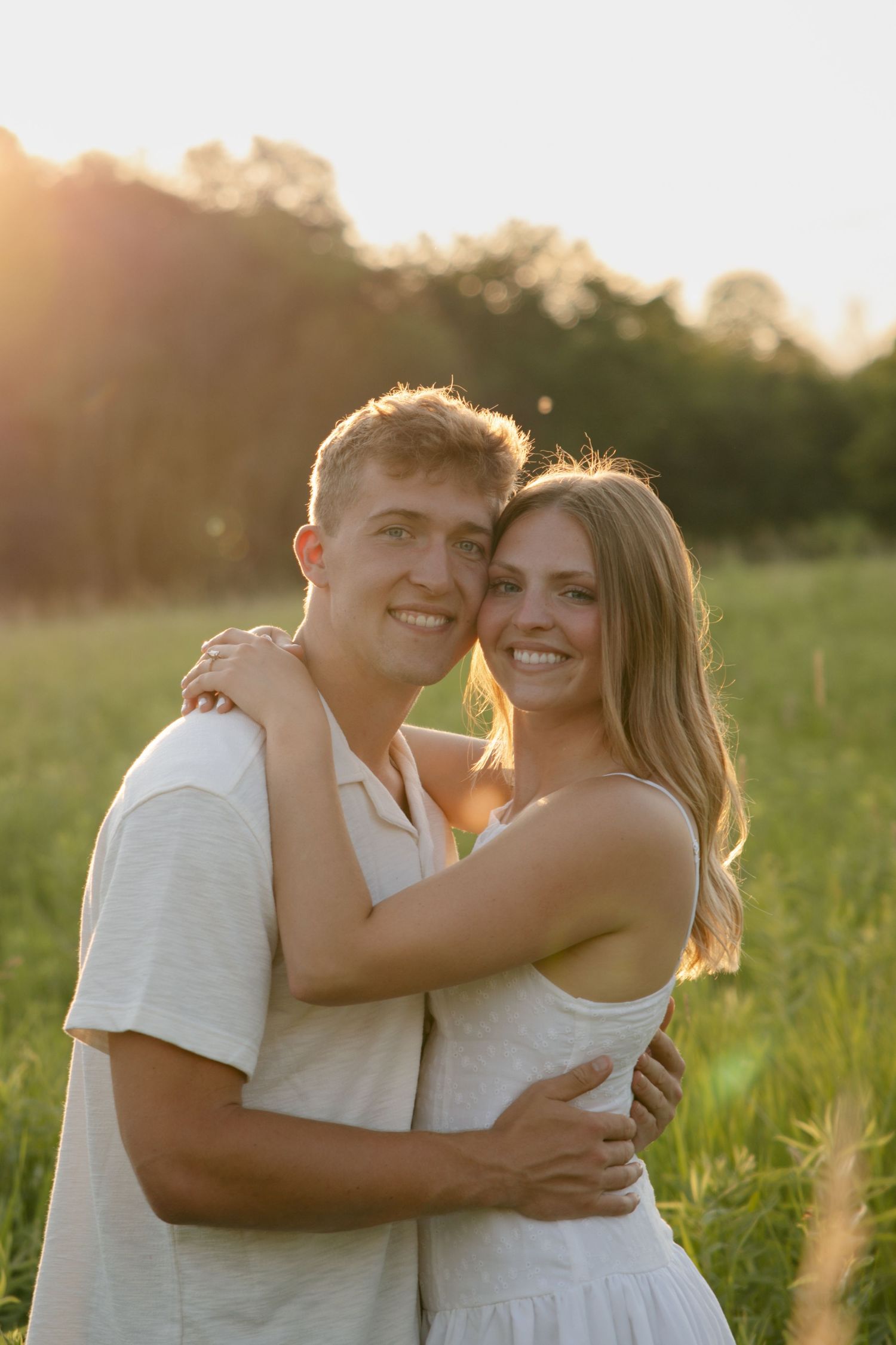 Smiling couple hugging closely in a green field bathed in warm golden sunset light.