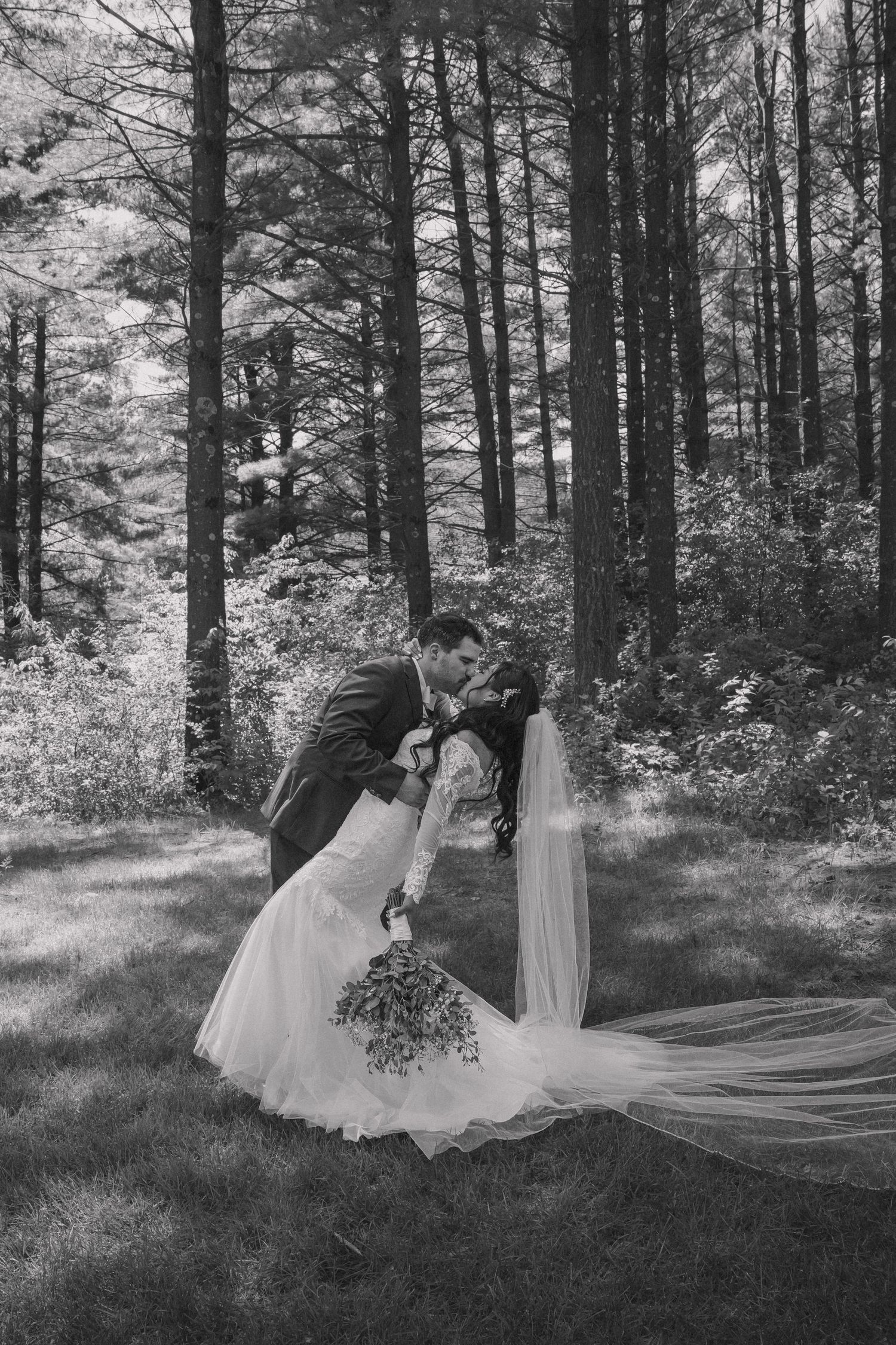 Black and white photo of a groom dipping and kissing his bride in a forest, her veil flowing gracefully.