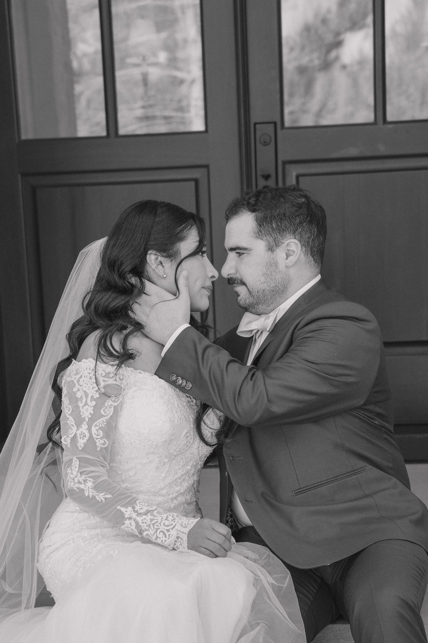 Black and white close-up of groom tenderly cupping brides face near elegant dark double doors.