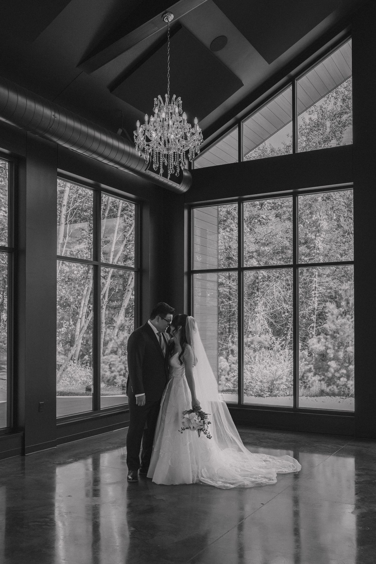 Black and white photo of bride and groom sharing intimate moment under crystal chandelier by large windows.