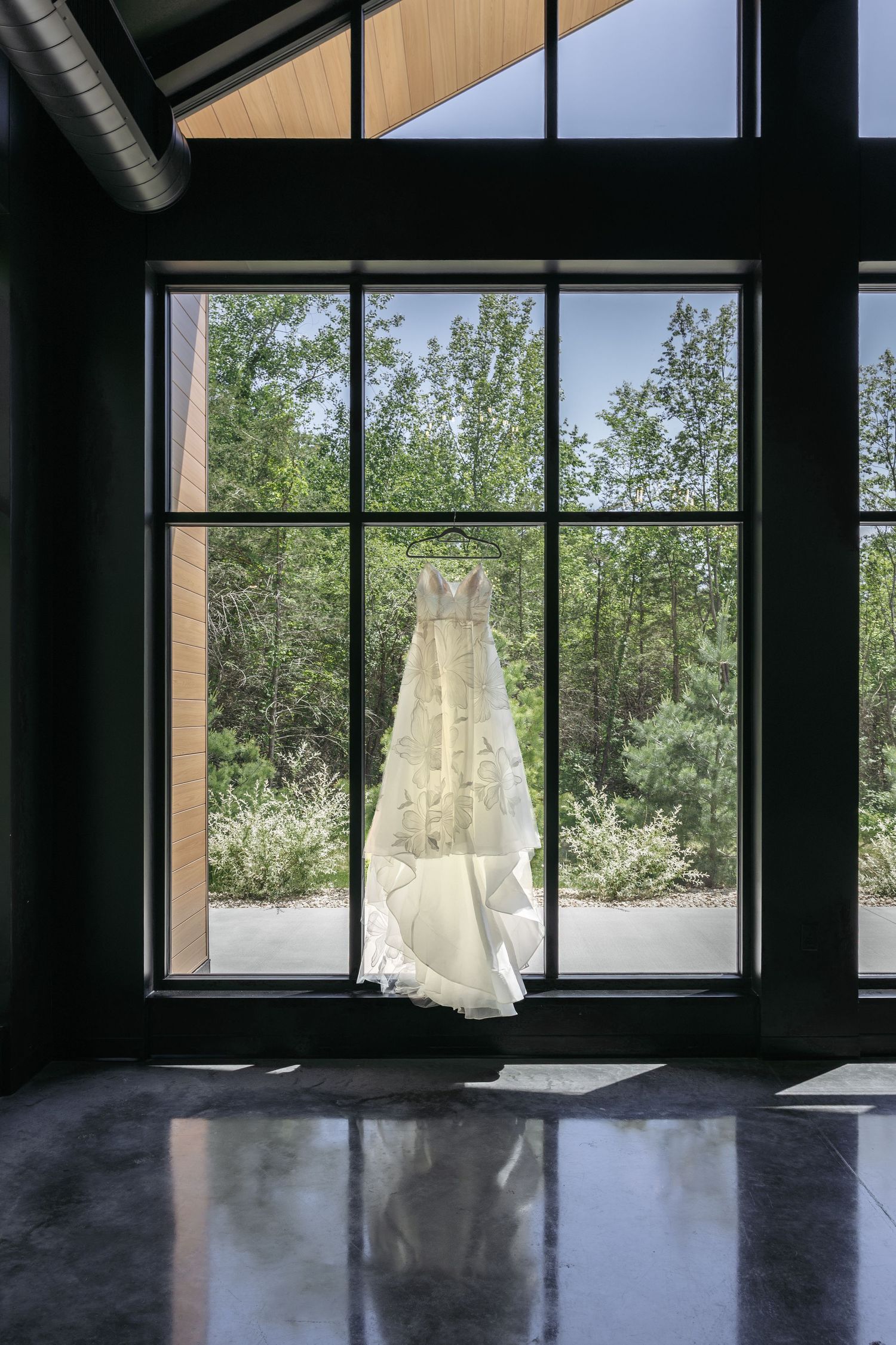 Stunning wedding dress hanging in a large black-framed window reflecting on polished concrete floors.