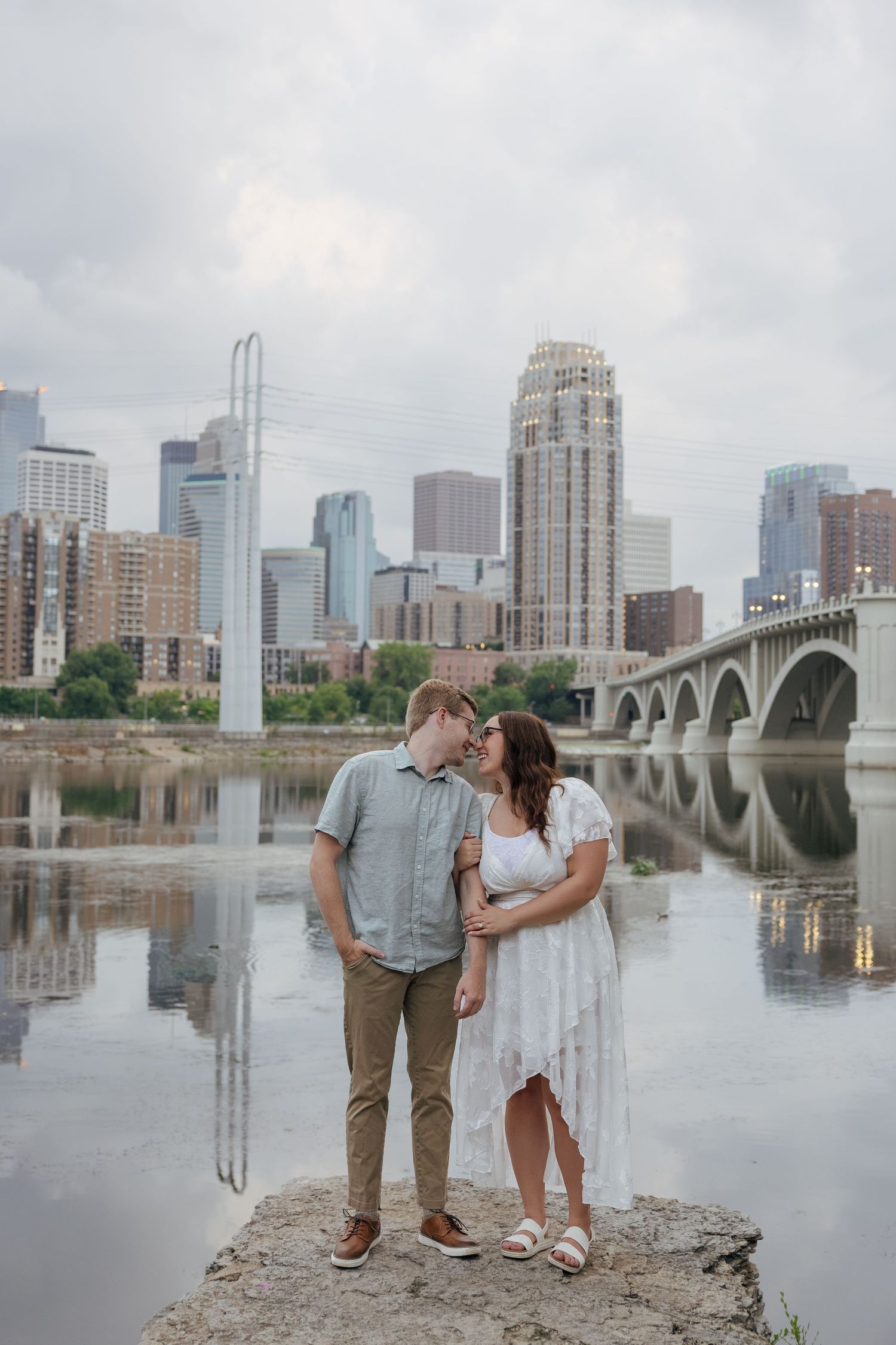 Couple shares a romantic moment on riverbank with Minneapolis skyline and bridge reflected in calm water behind them.