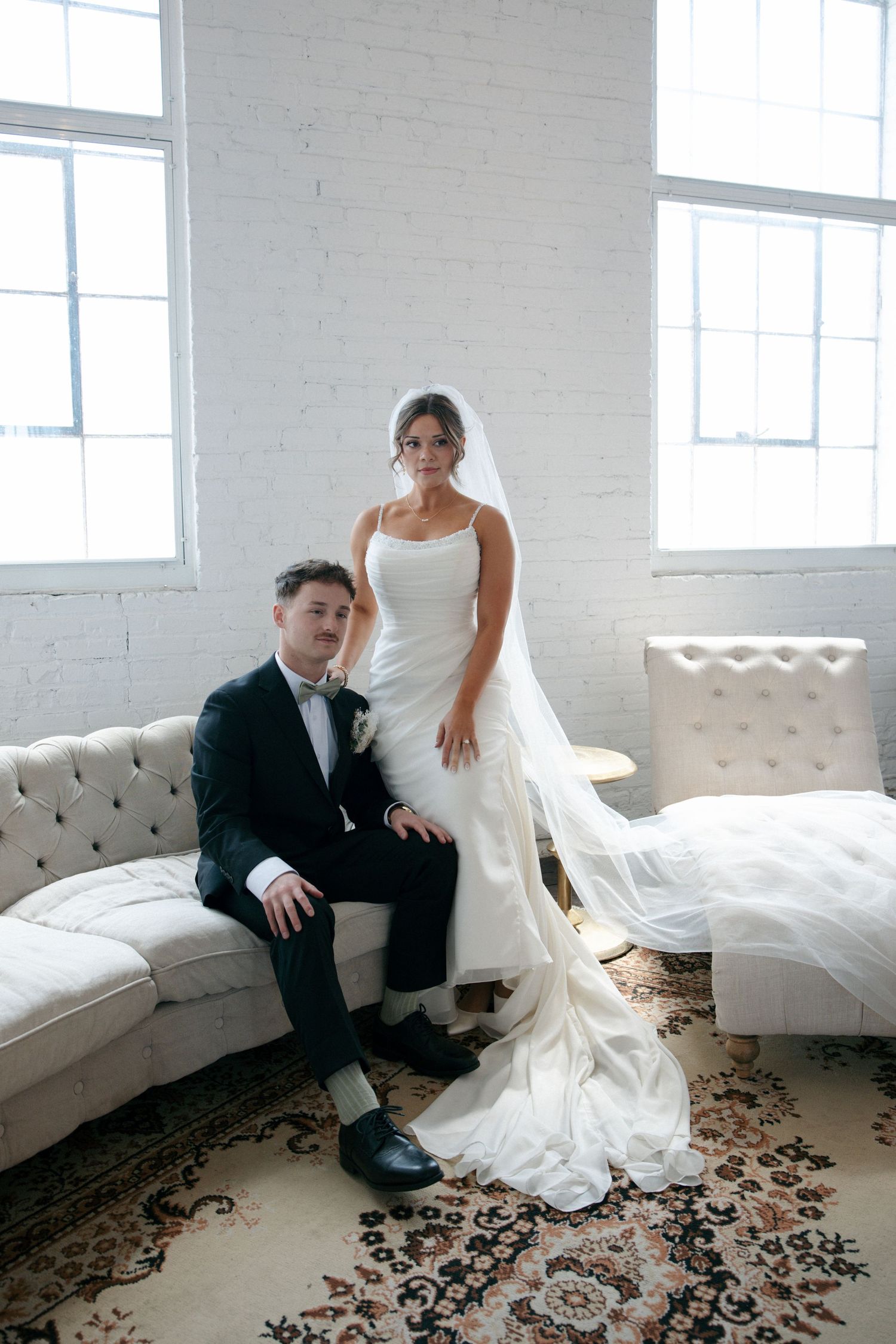 Bride in white gown and veil stands beside groom in black tuxedo seated on tufted sofa in bright white brick venue.