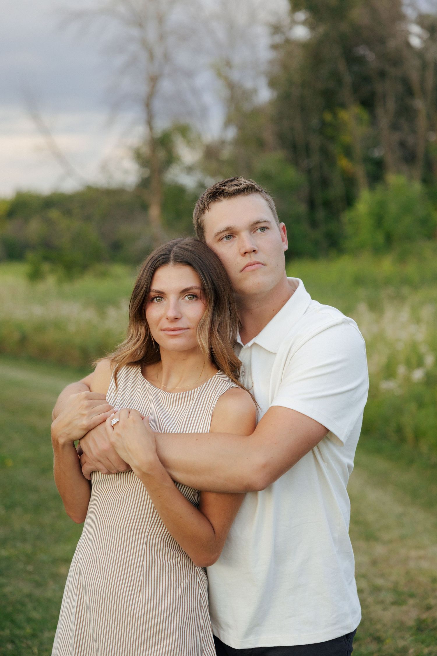 Couple posing seriously for portrait outdoors, man embracing woman from behind near green trees.