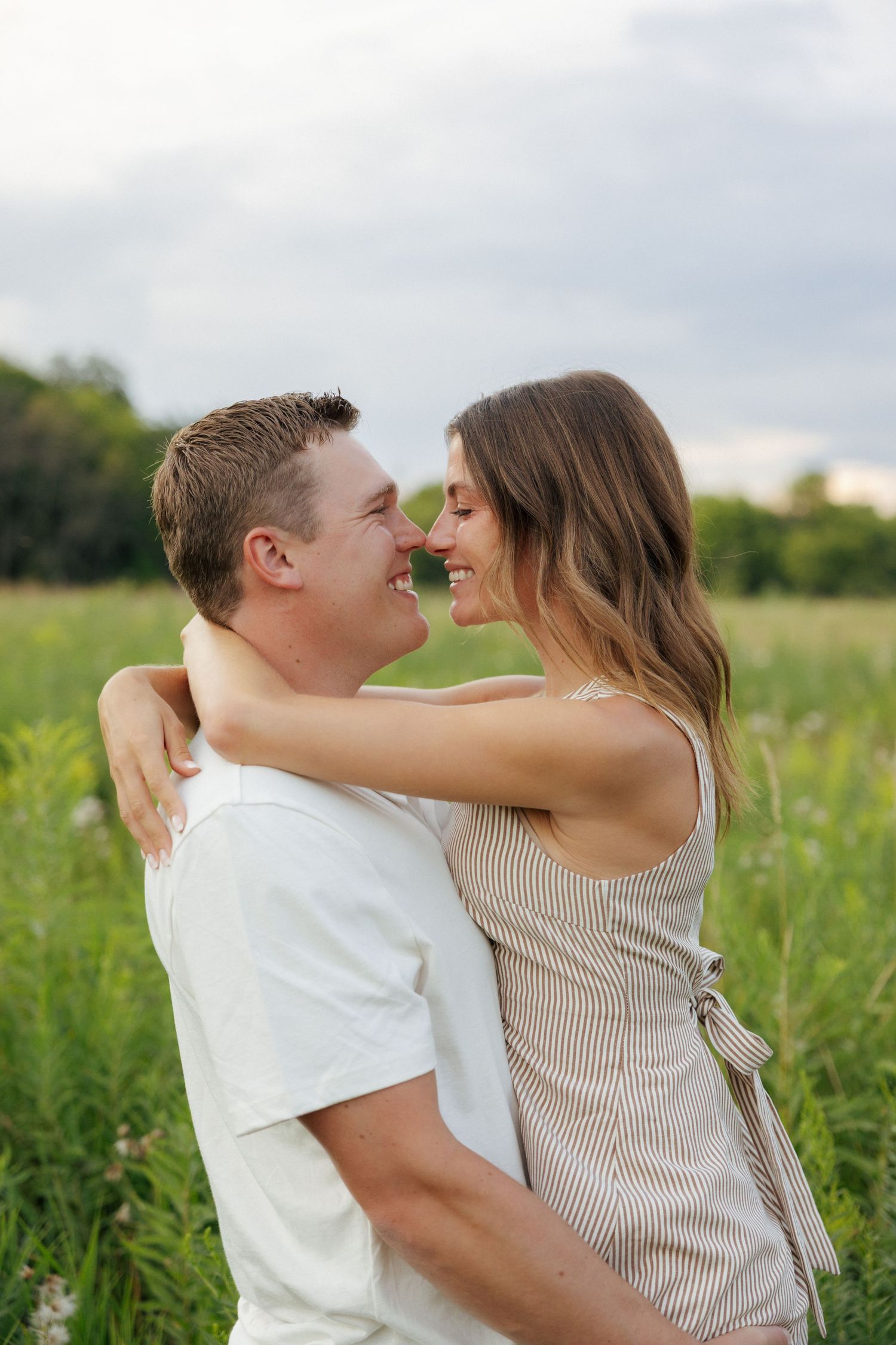 Couple embracing and smiling at each other in a lush green field with wildflowers under a cloudy sky.