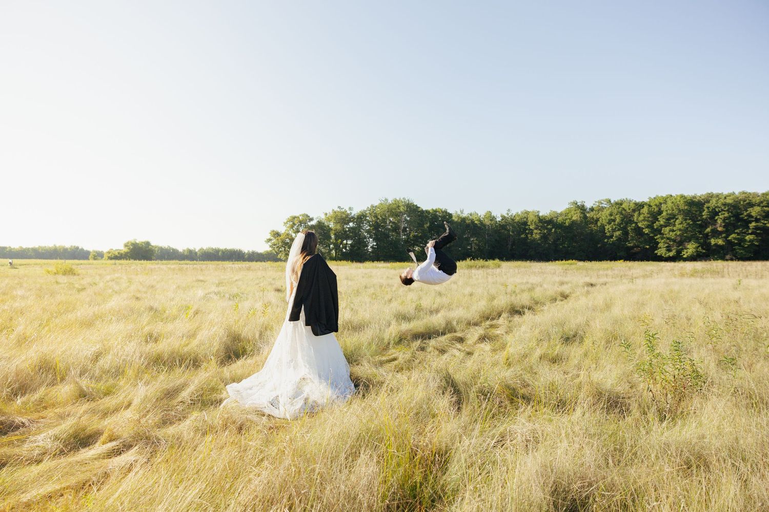 Bride and groom walking through a golden sunlit field with trees in the background on their wedding day.