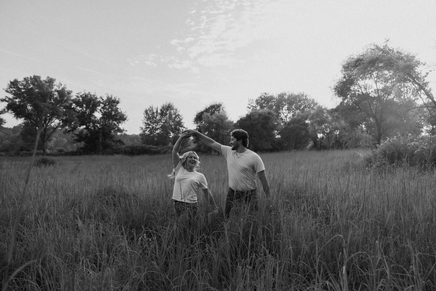 Black and white photo of two people holding hands in a tall grass field with trees in the background.