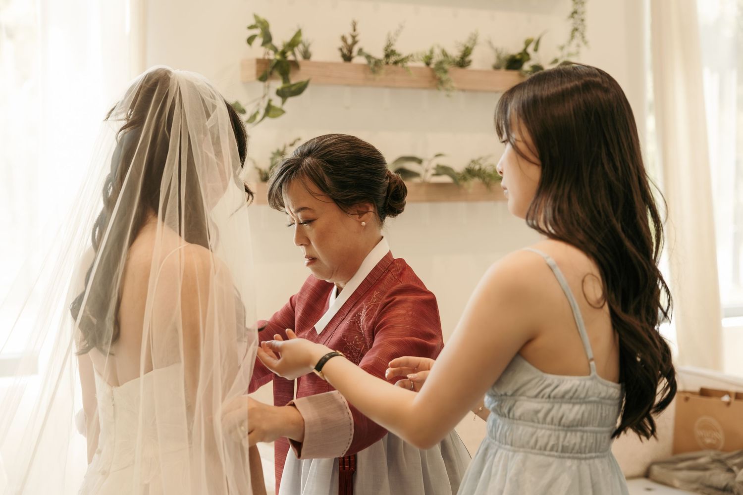 A bride in a white veil gets assistance from two women in a bright, elegant room during wedding preparations.