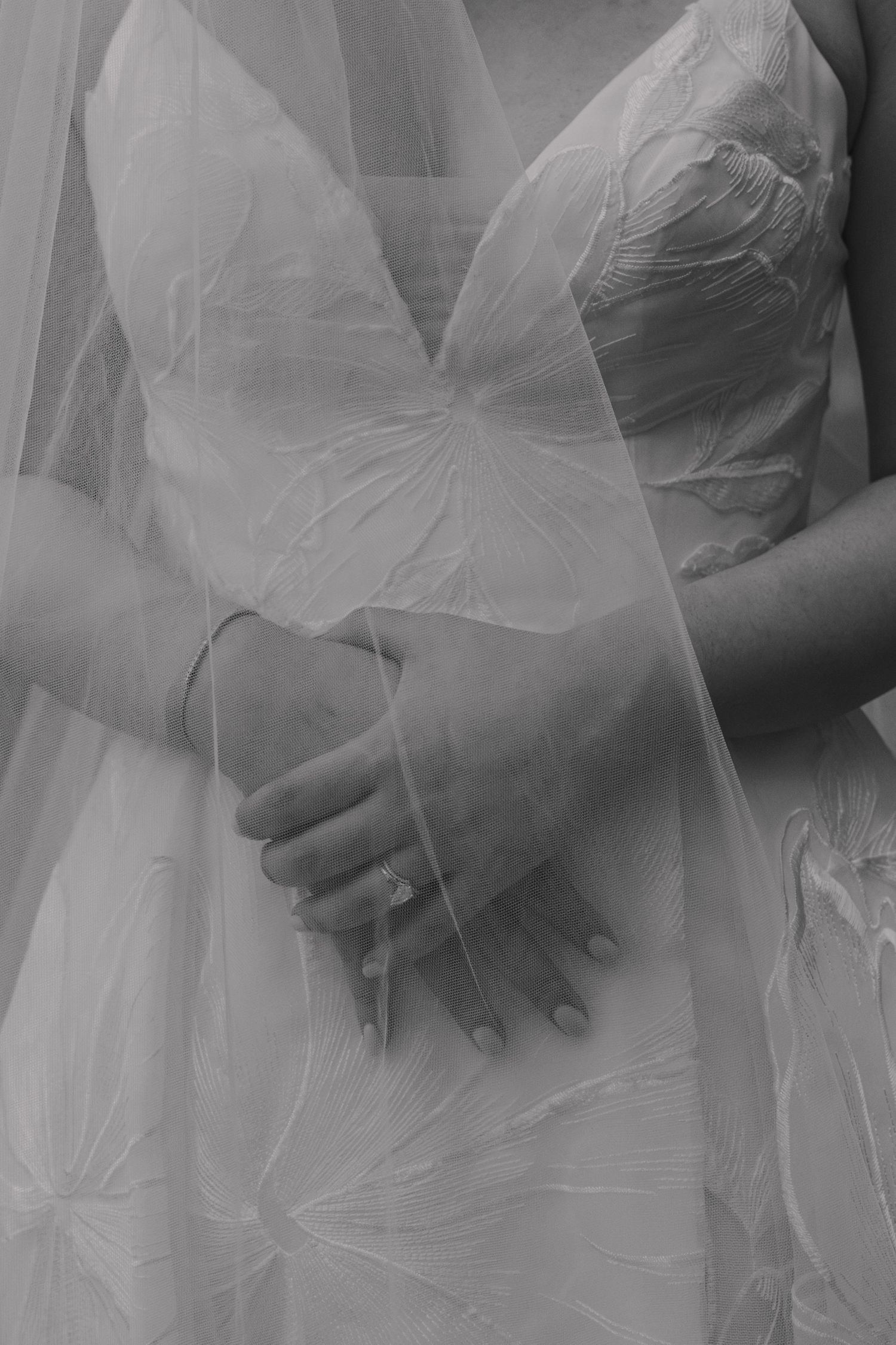 Black and white photo of a bride clasping hands over her wedding gown, showcasing an elegant ring and delicate veil.