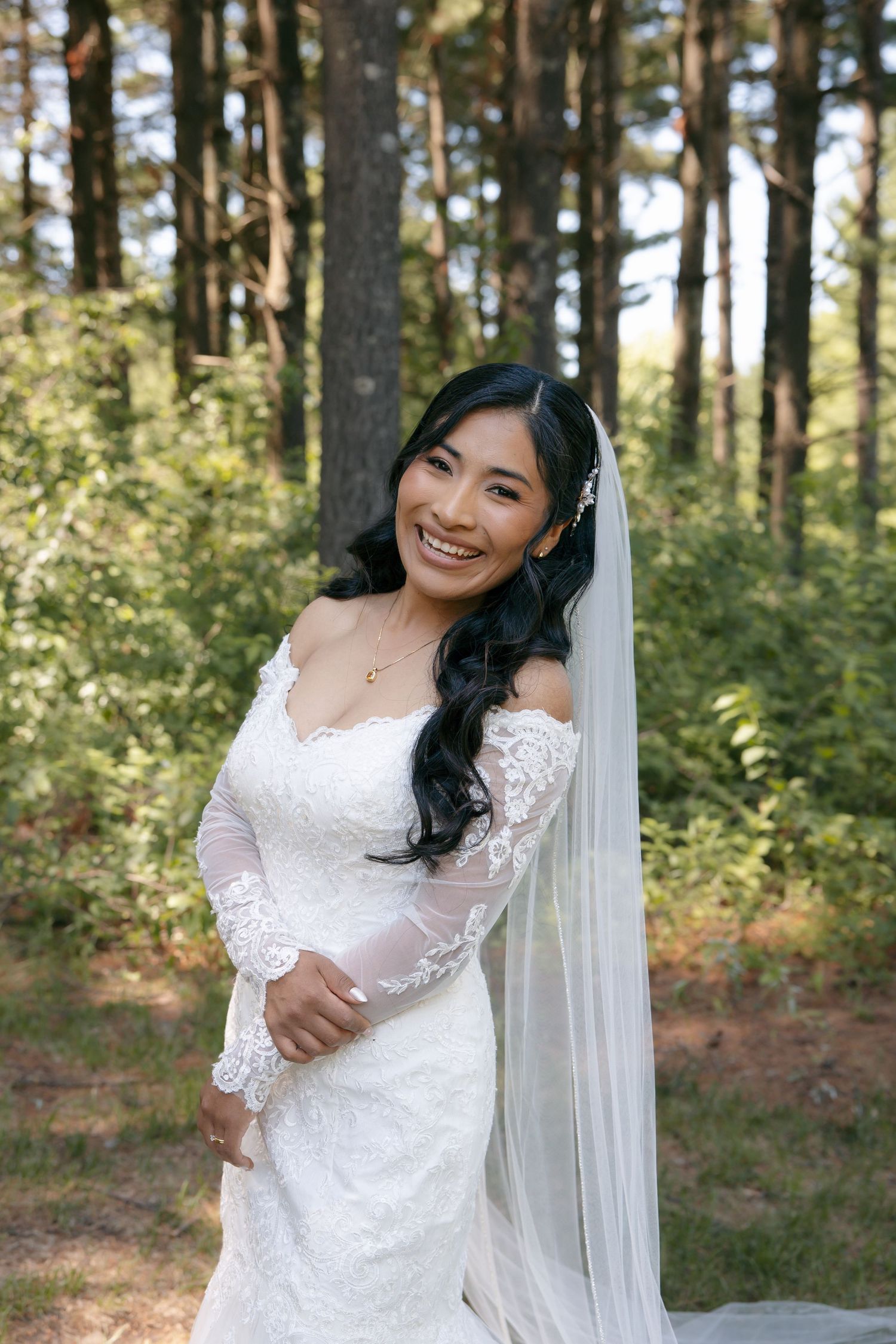 Smiling bride in lace off-shoulder gown and veil poses in sunlit forest.