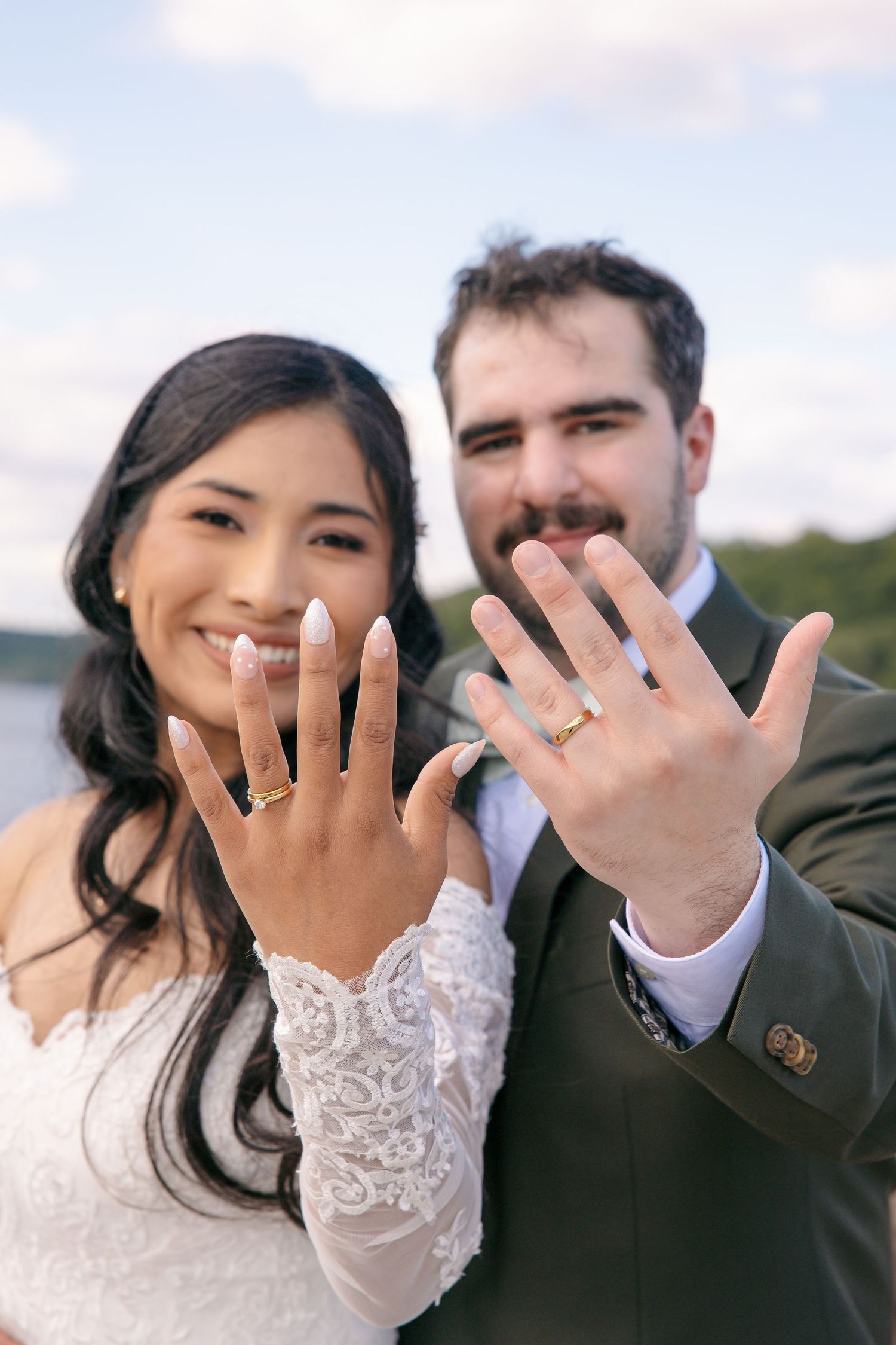 Newlywed couple smiling and showing off their wedding rings to the camera with a lake backdrop.
