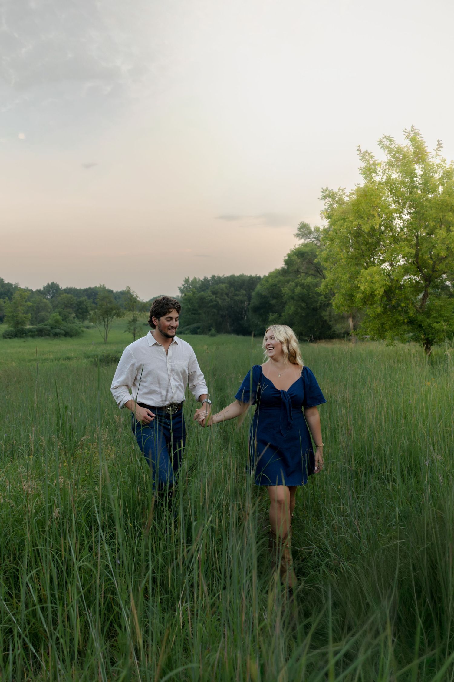 Couple holding hands walking through tall green grass field at sunset, man in white shirt and woman in navy dress.