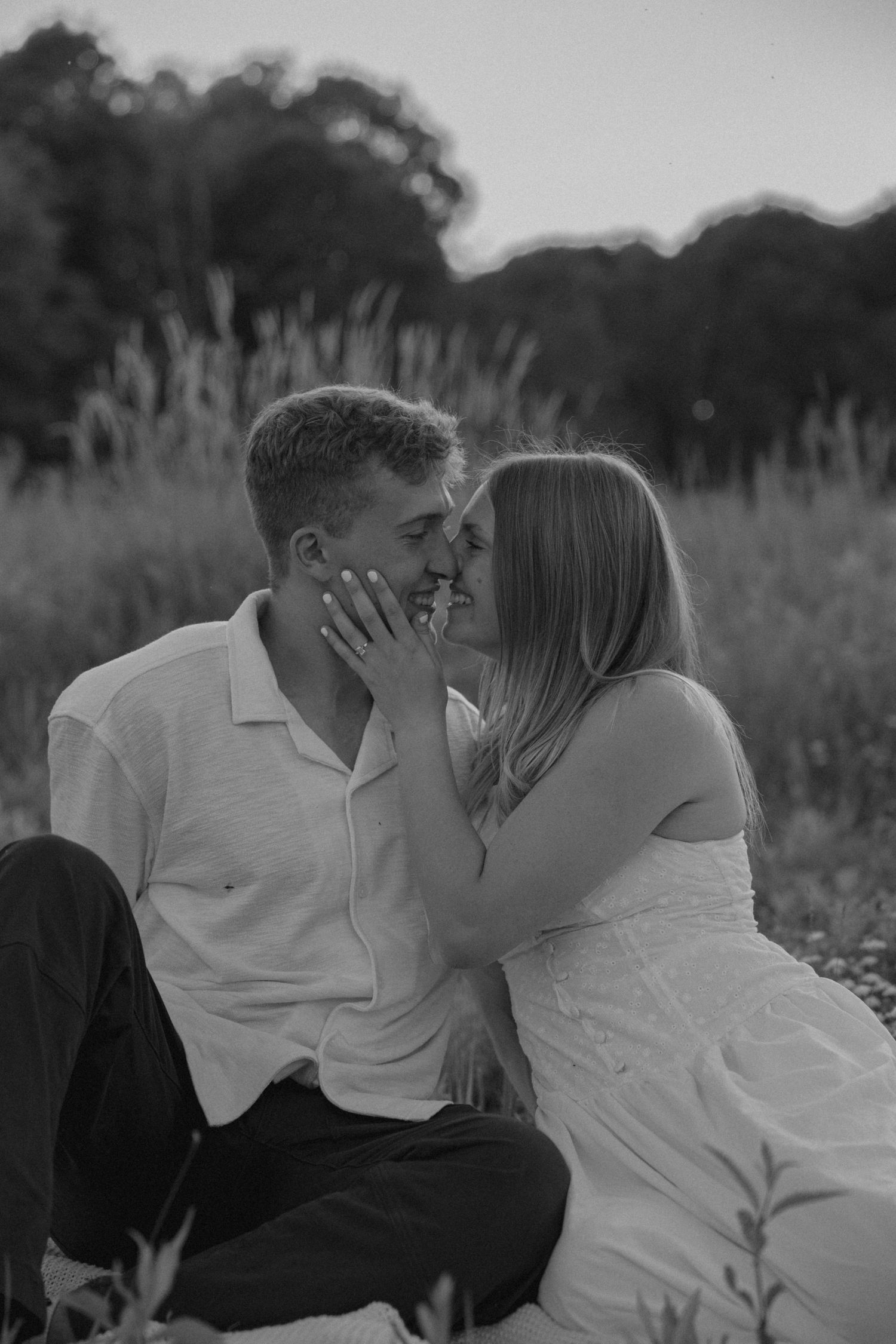 Black and white photo of a couple sharing a tender kiss while sitting in a meadow at sunset.