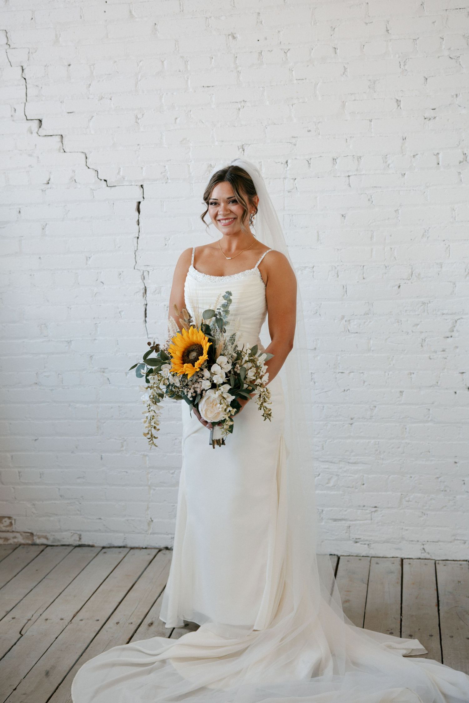 Smiling bride in white gown holding sunflower bouquet against white brick wall.