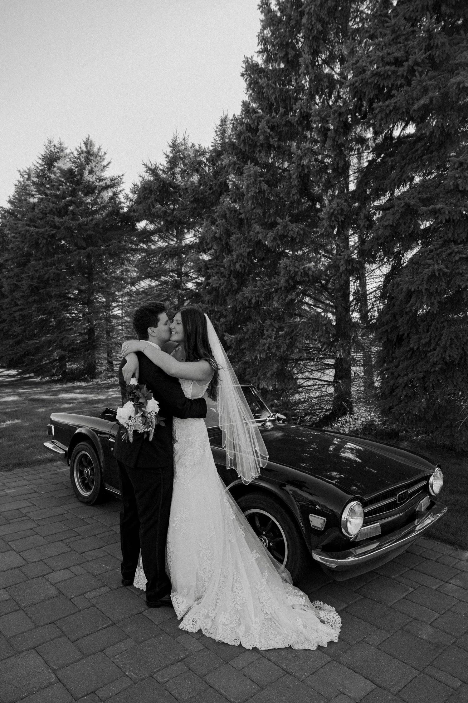 Black and white photo of bride and groom kissing in front of a classic vintage car surrounded by tall evergreen trees.