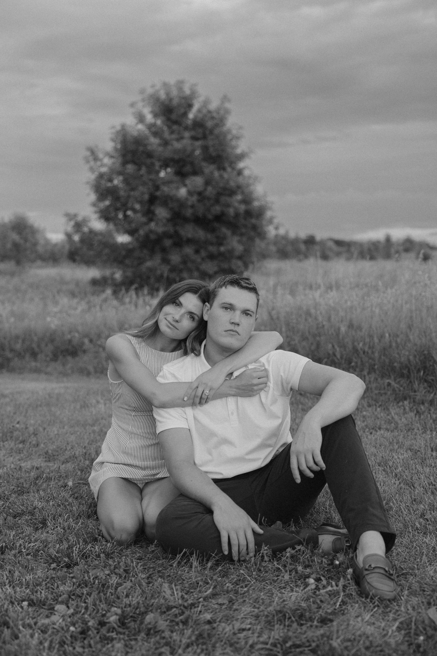 Black and white photo of a young couple sitting on grass in a field, woman embracing man from behind near a tree.