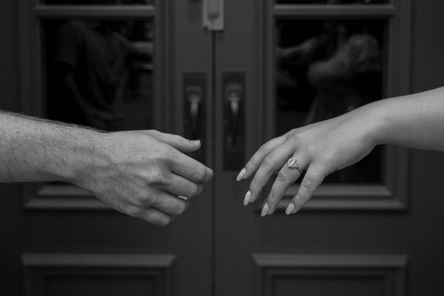 Black and white photo of two hands reaching toward each other, woman wearing engagement ring, near dark wooden doors.