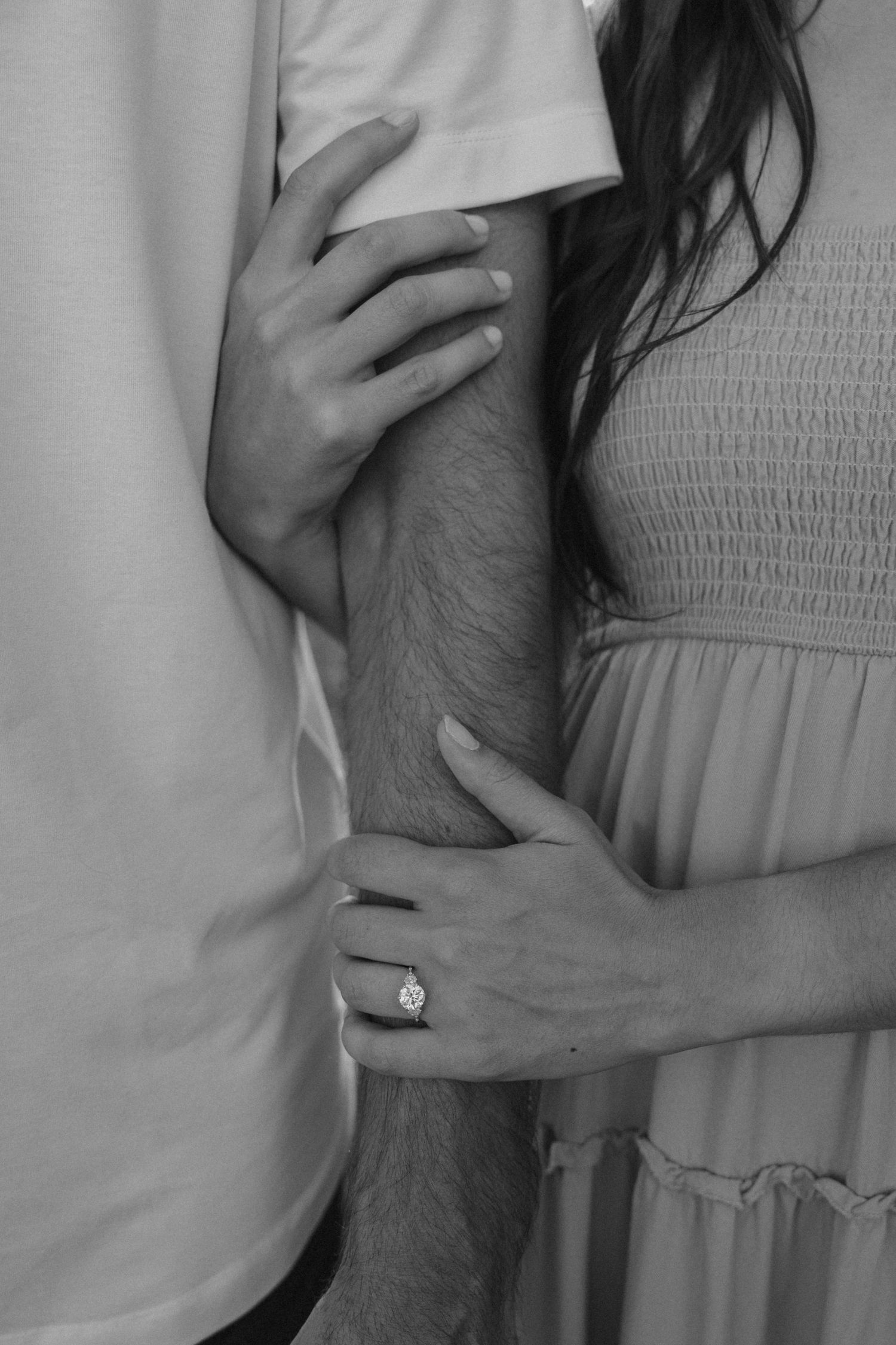 Black and white photo of a couple embracing, woman wearing an engagement ring holding mans arm in a romantic pose.