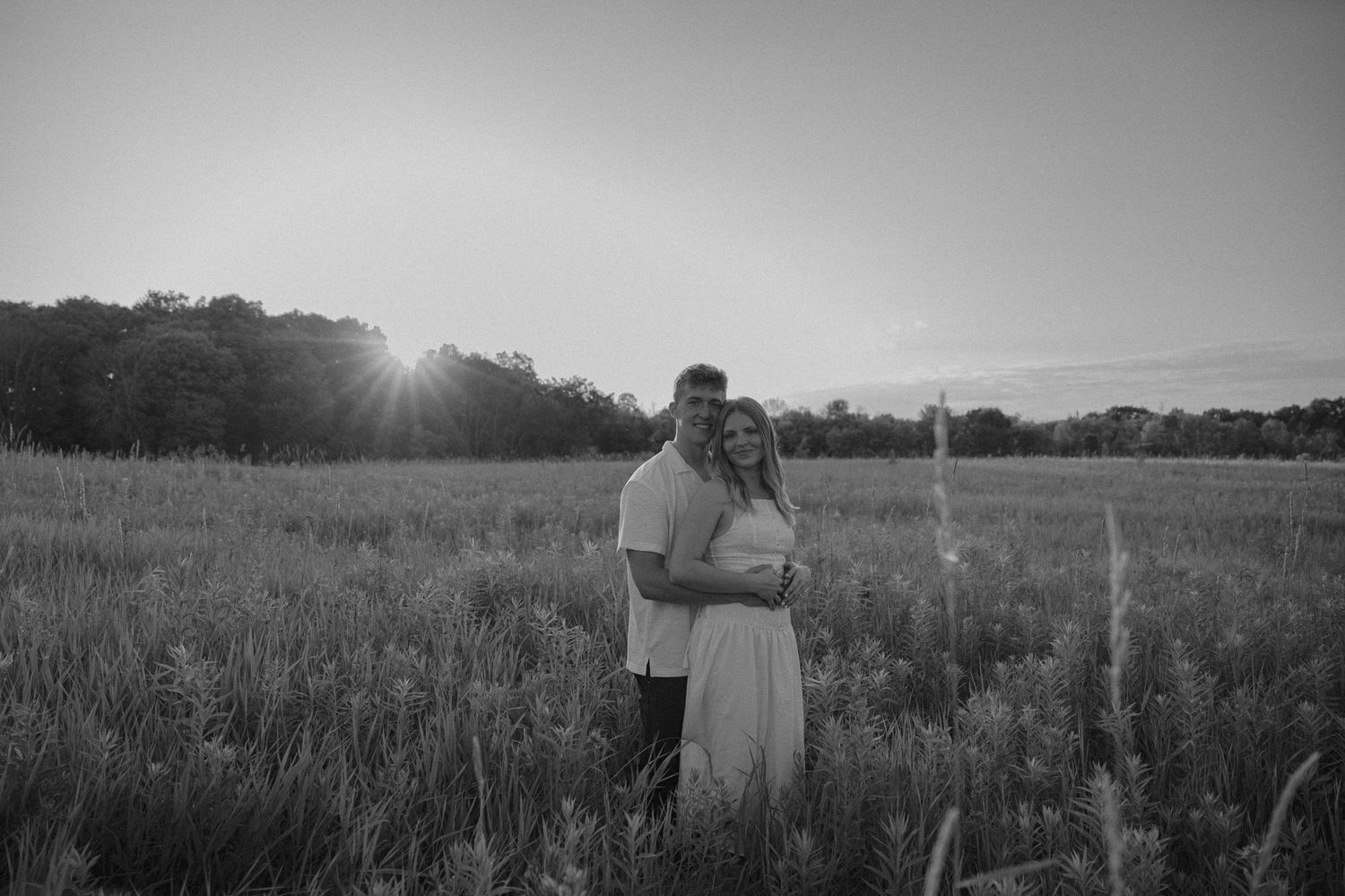 Black and white photo of a couple embracing in a sunlit field at sunset with trees in the background.