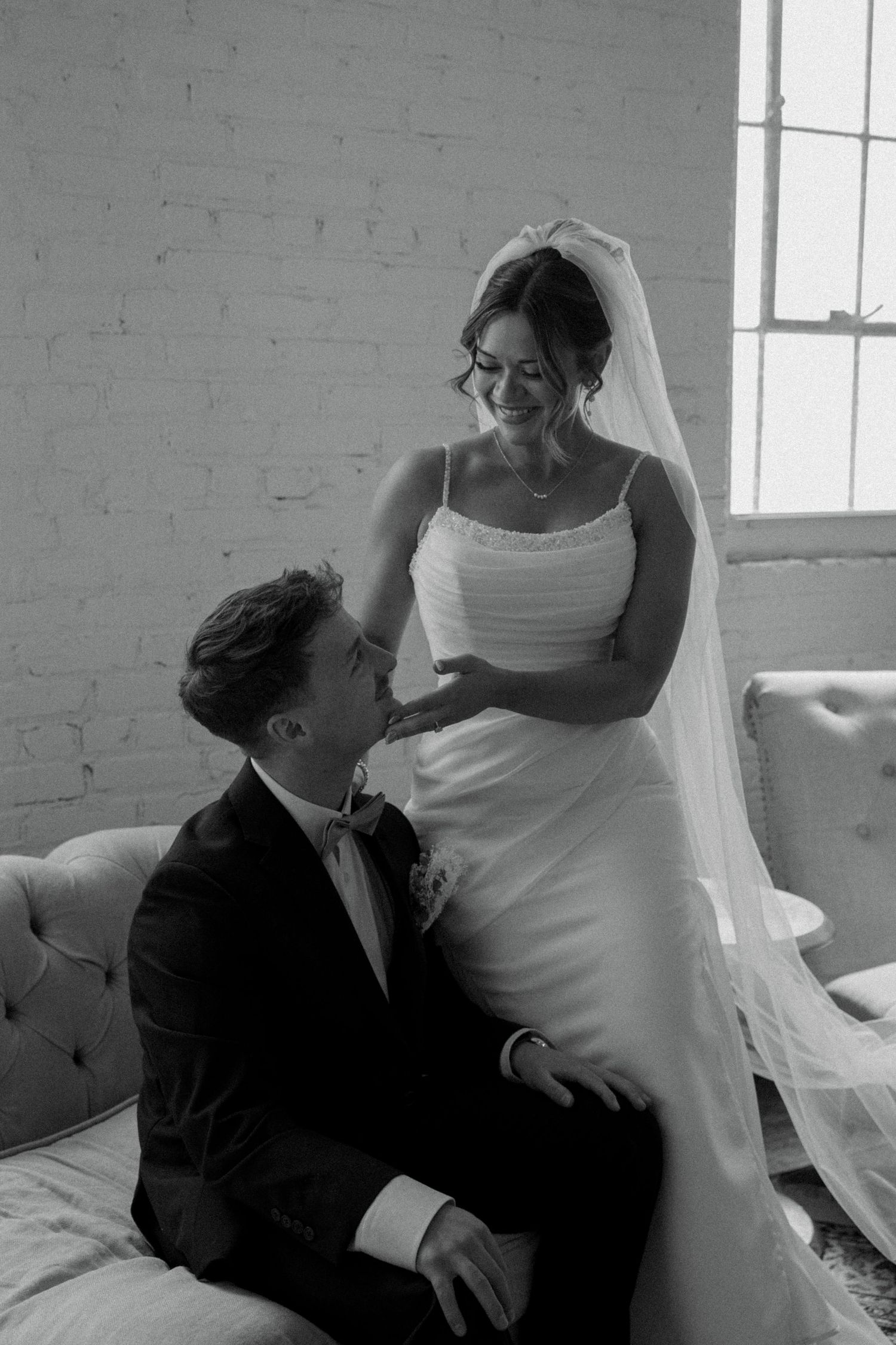Black and white photo of a smiling bride in a veil standing over a seated groom in a tuxedo on a couch.
