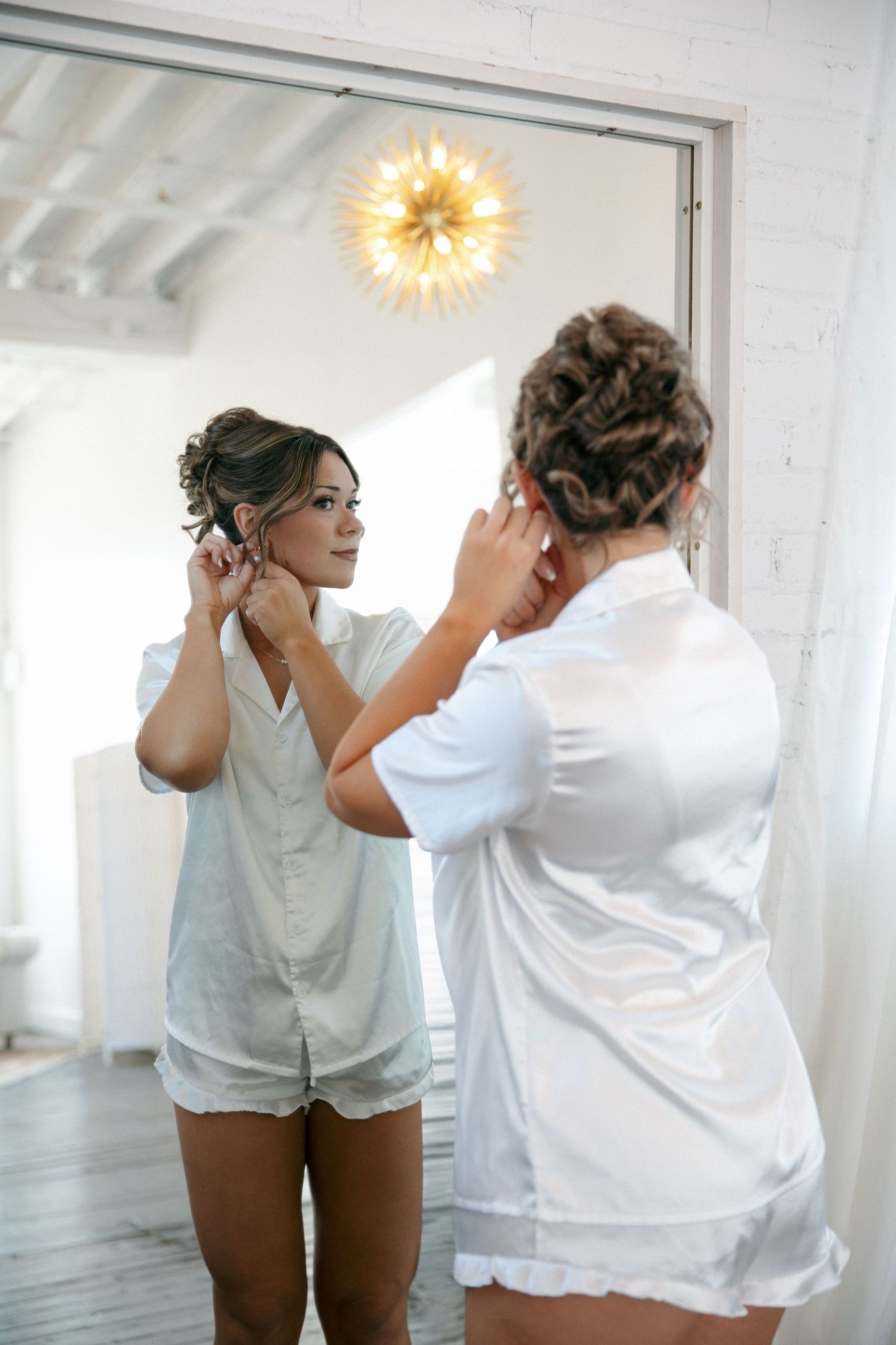 Bride in white satin robe putting on earrings while looking in a large mirror during wedding morning prep.