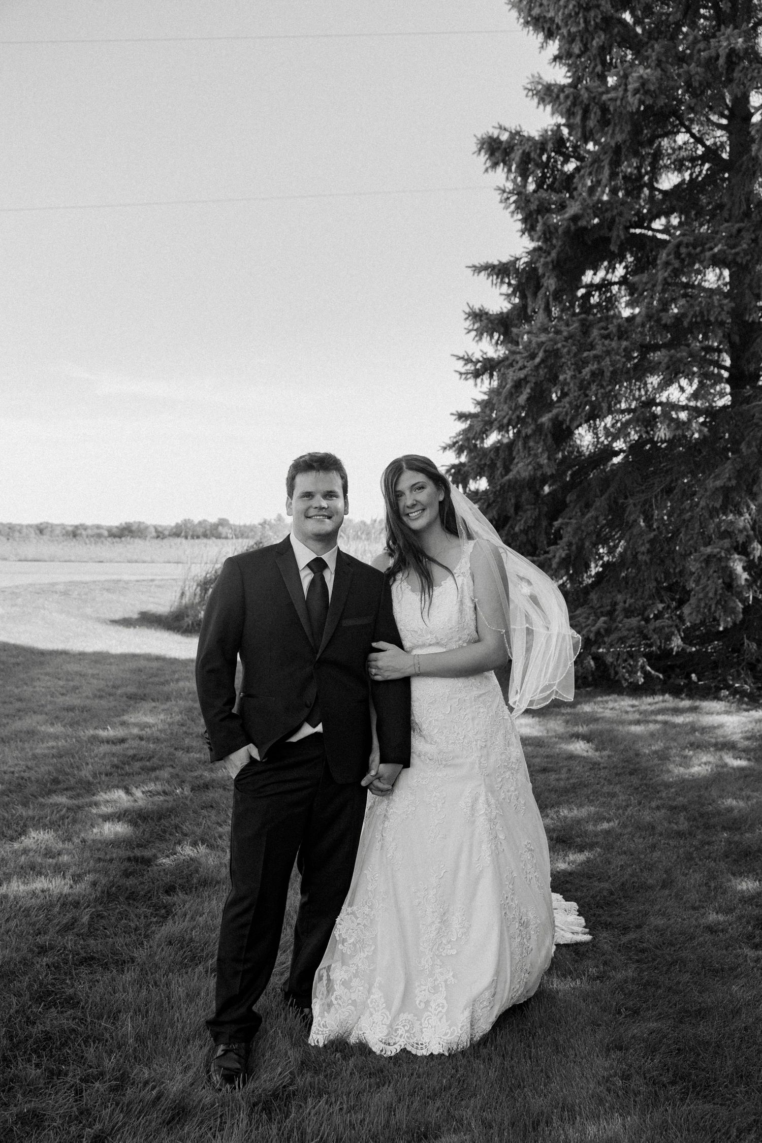 Black and white portrait of smiling bride and groom standing together outdoors near evergreen tree.