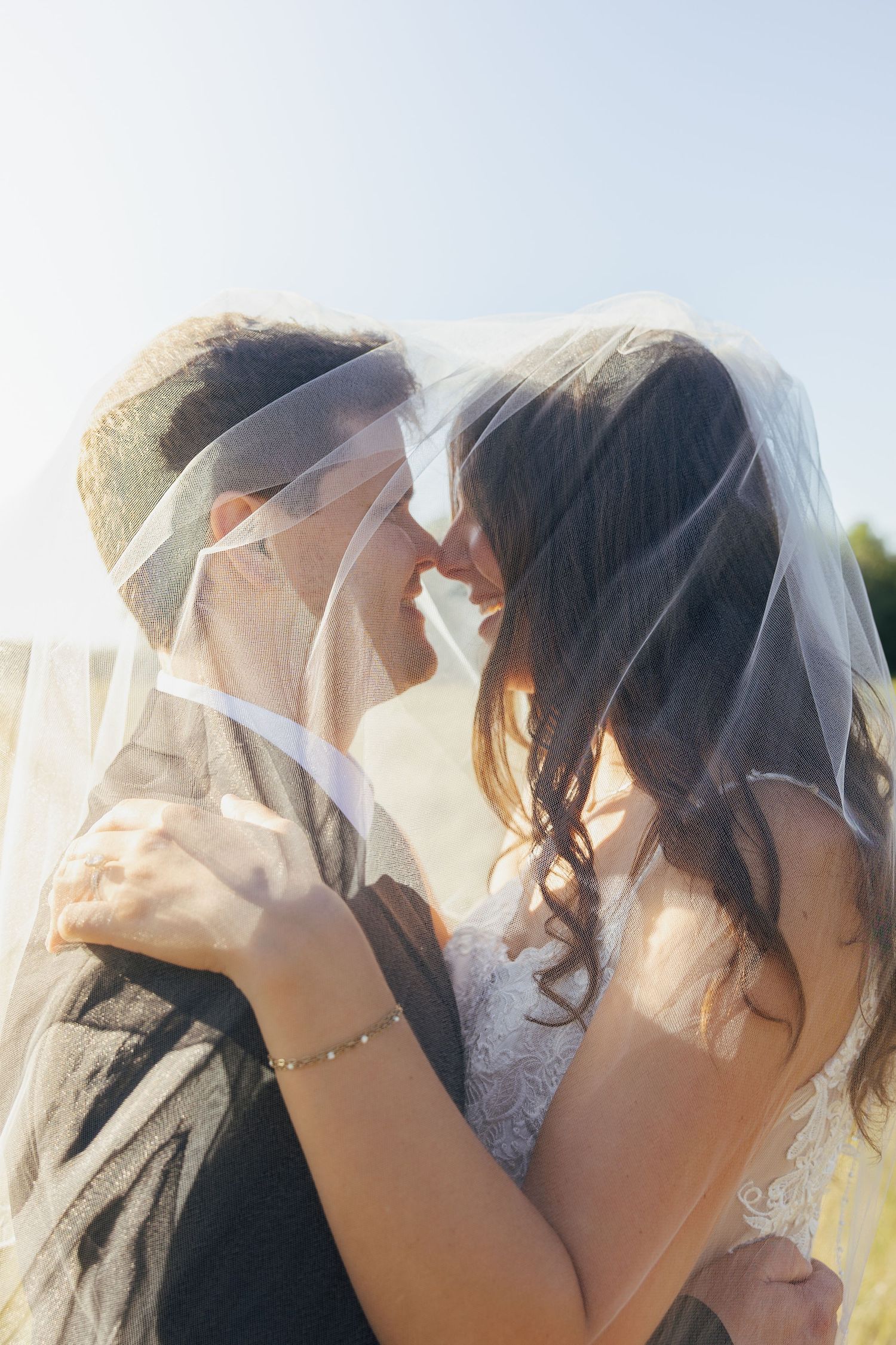 Bride and groom smiling closely together under sheer wedding veil in bright sunlight.