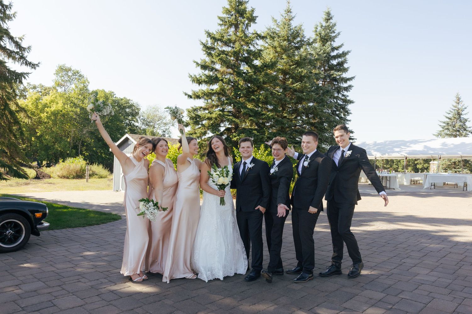 Wedding party poses outdoors near tall pine trees, bridesmaids in blush gowns and groomsmen in black suits.