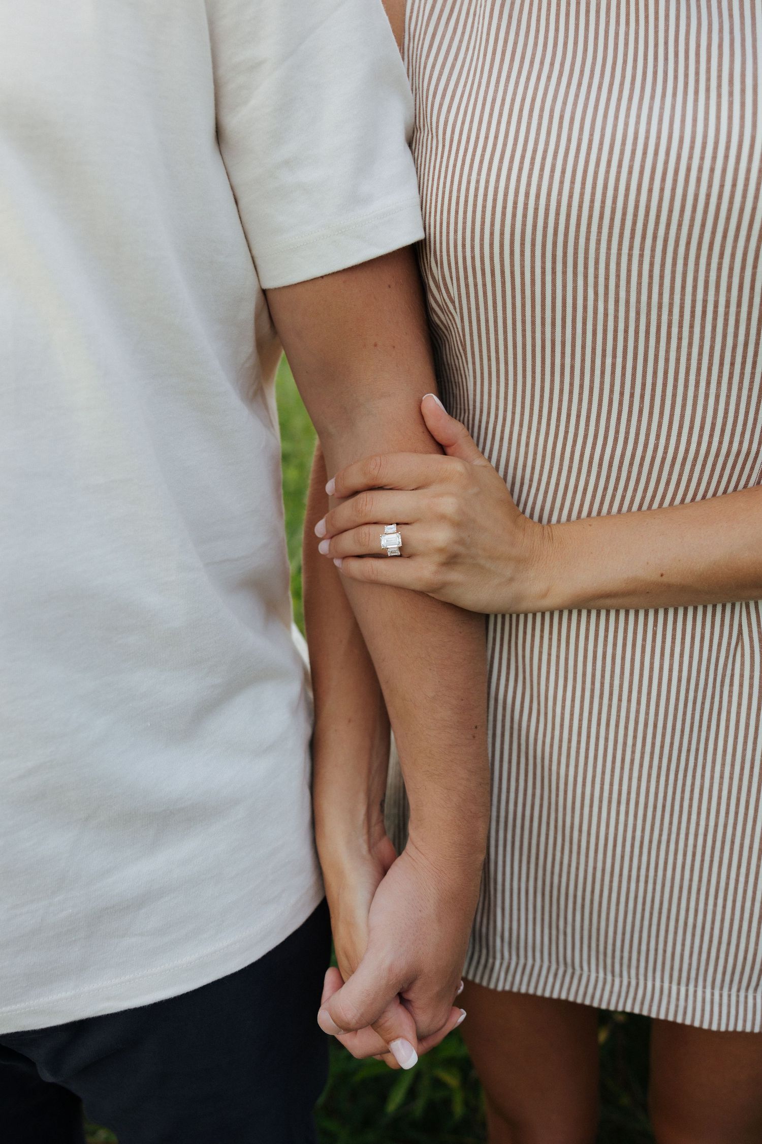 Couple holding hands with an engagement ring visible on the woman's finger, wearing a striped dress.