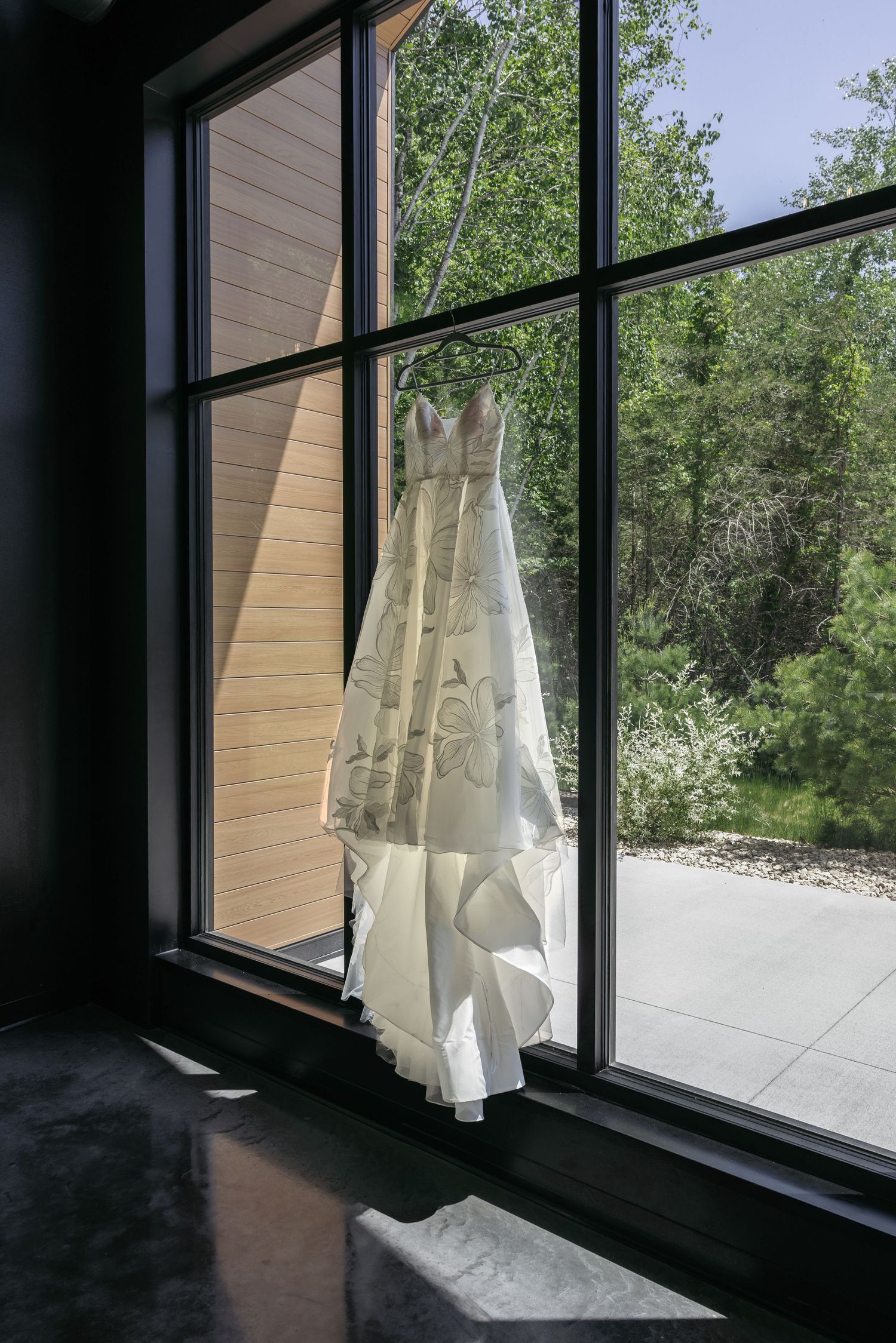Elegant lace wedding gown hanging in a sunlit window with a view of lush green trees outside.