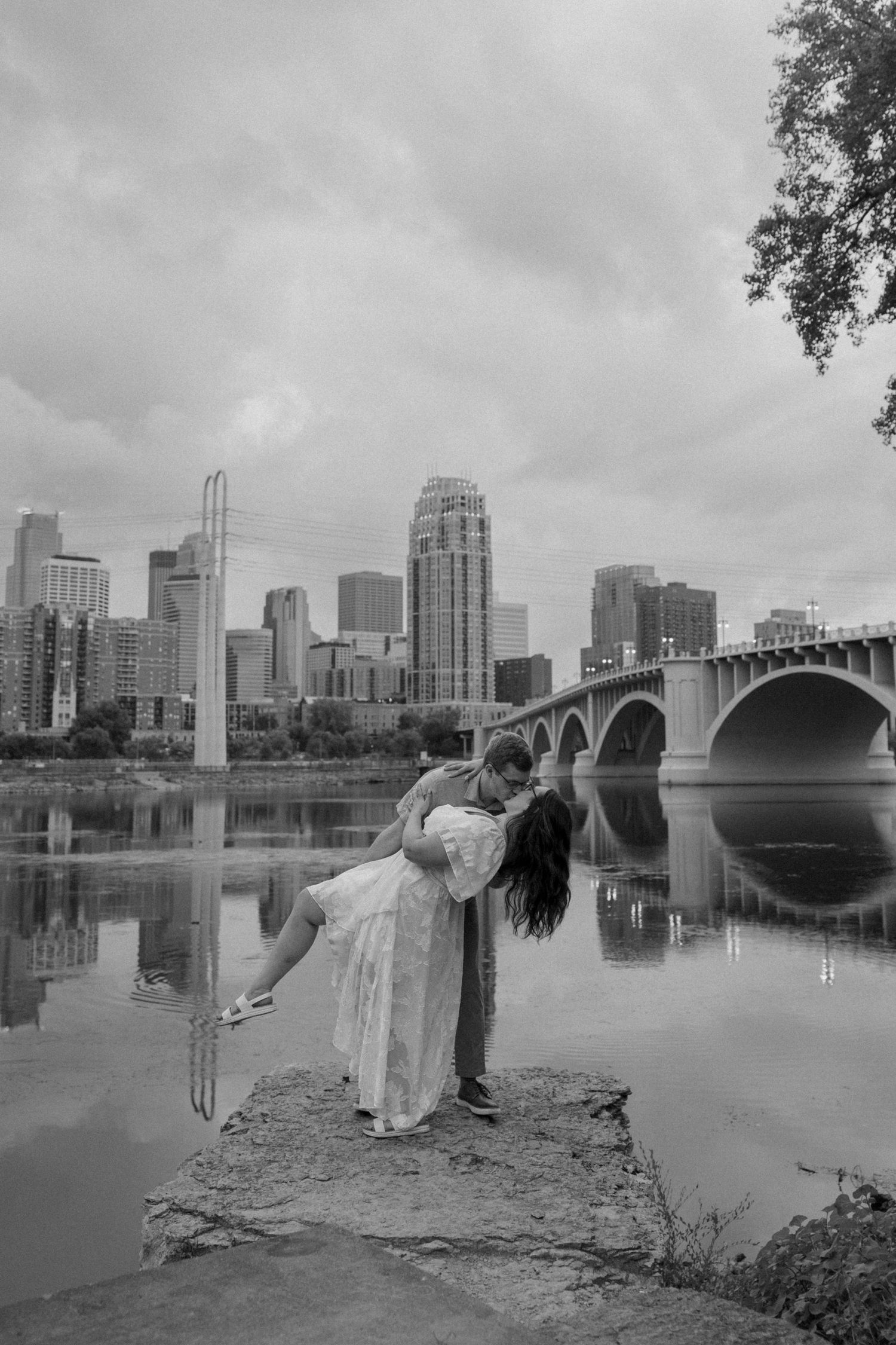Black and white photo of a couple embracing on rocks by a river with a city skyline and bridge in the background.