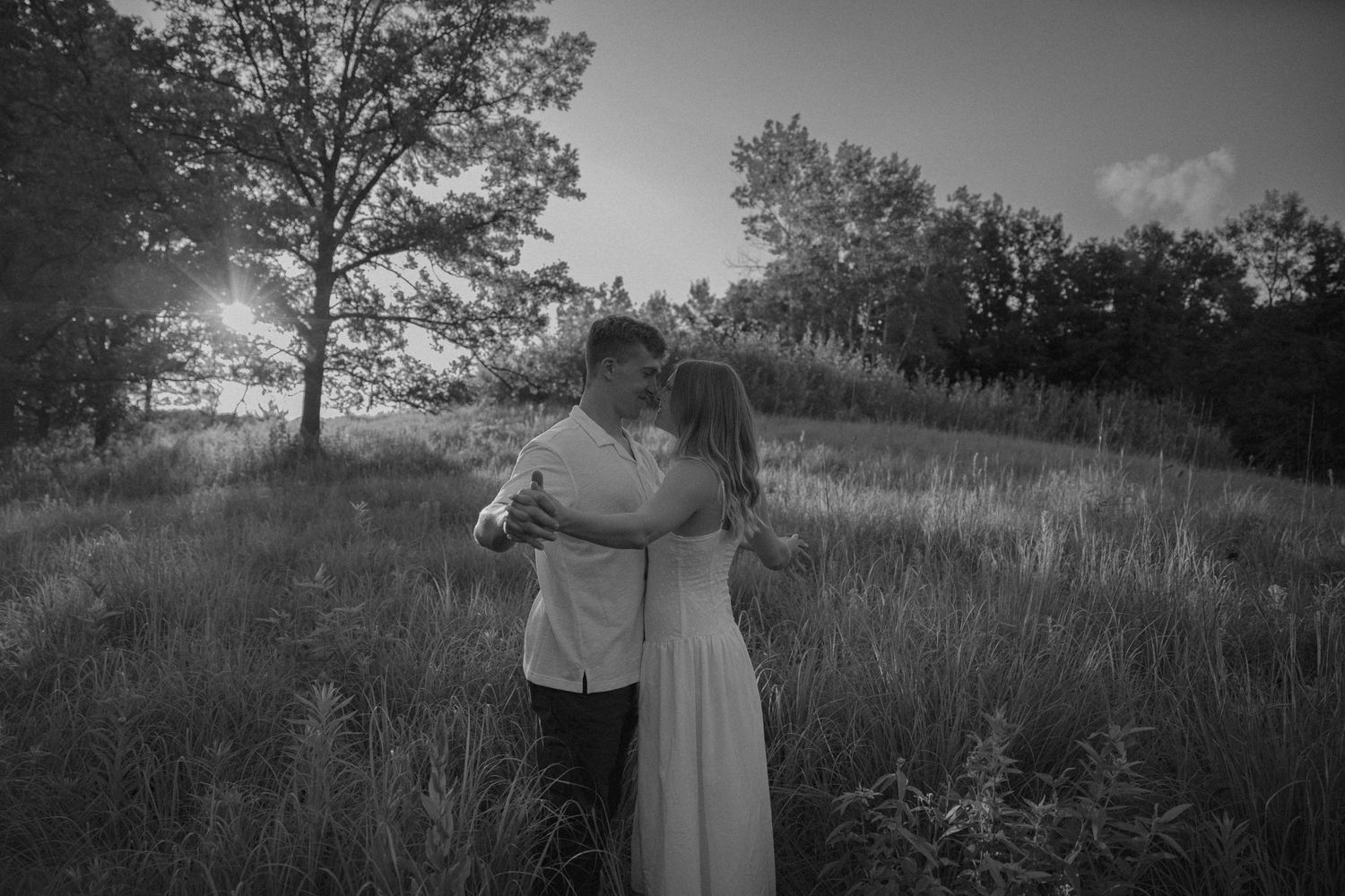 Romantic black and white photo of couple embracing in sunlit meadow surrounded by tall grass and trees.