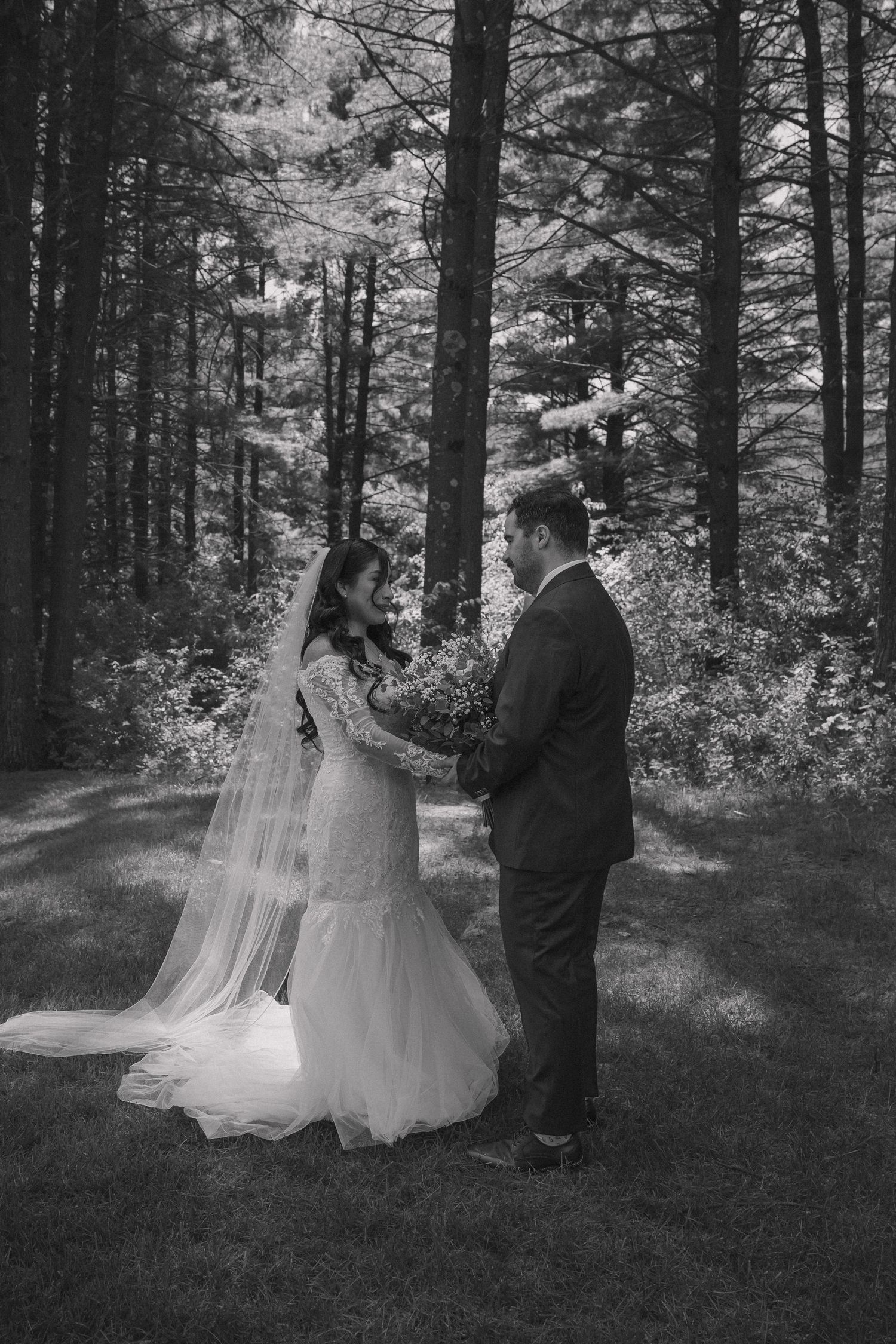 Black and white photo of bride and groom holding hands facing each other in forest.