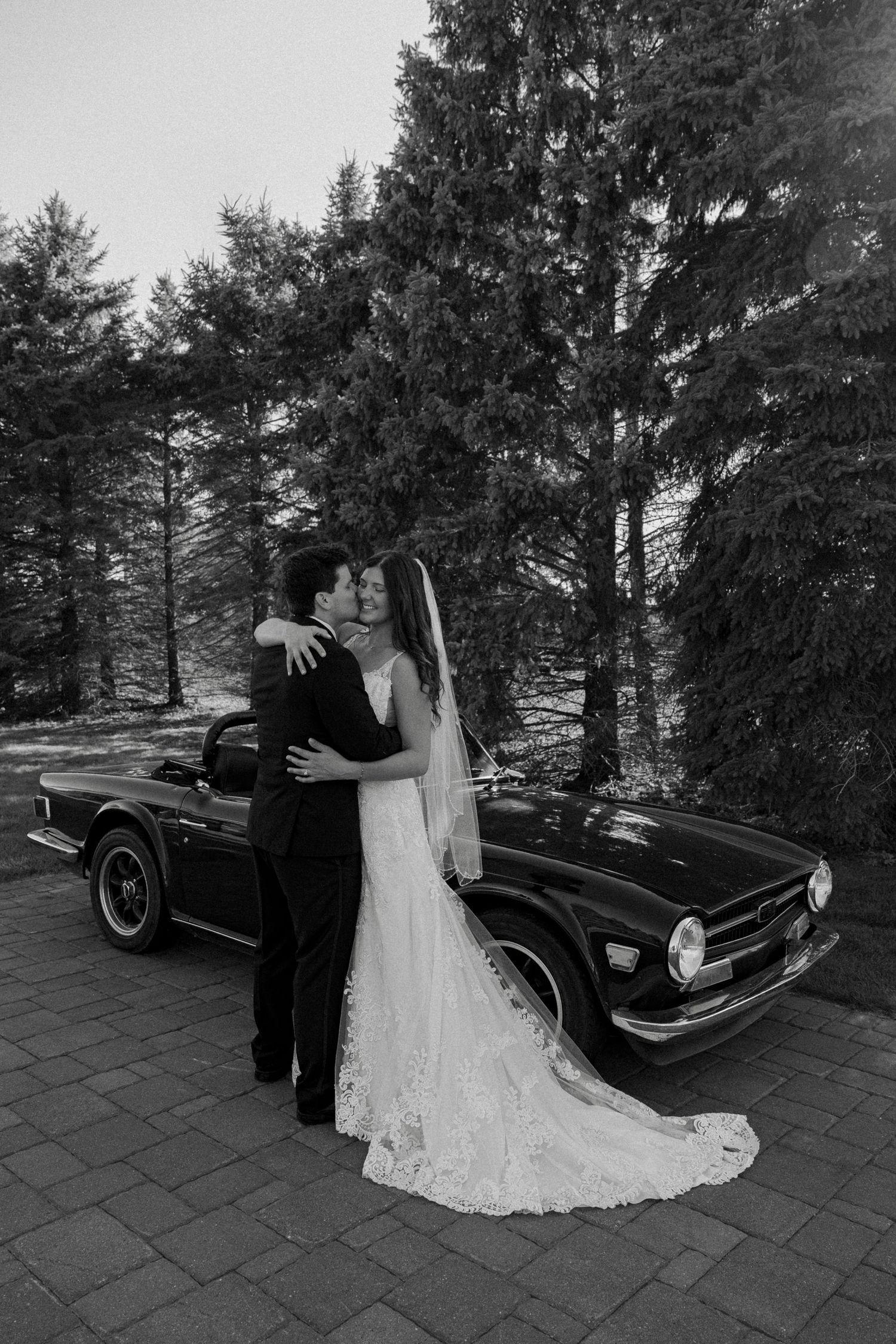 Black and white photo of bride and groom kissing in front of vintage classic car.