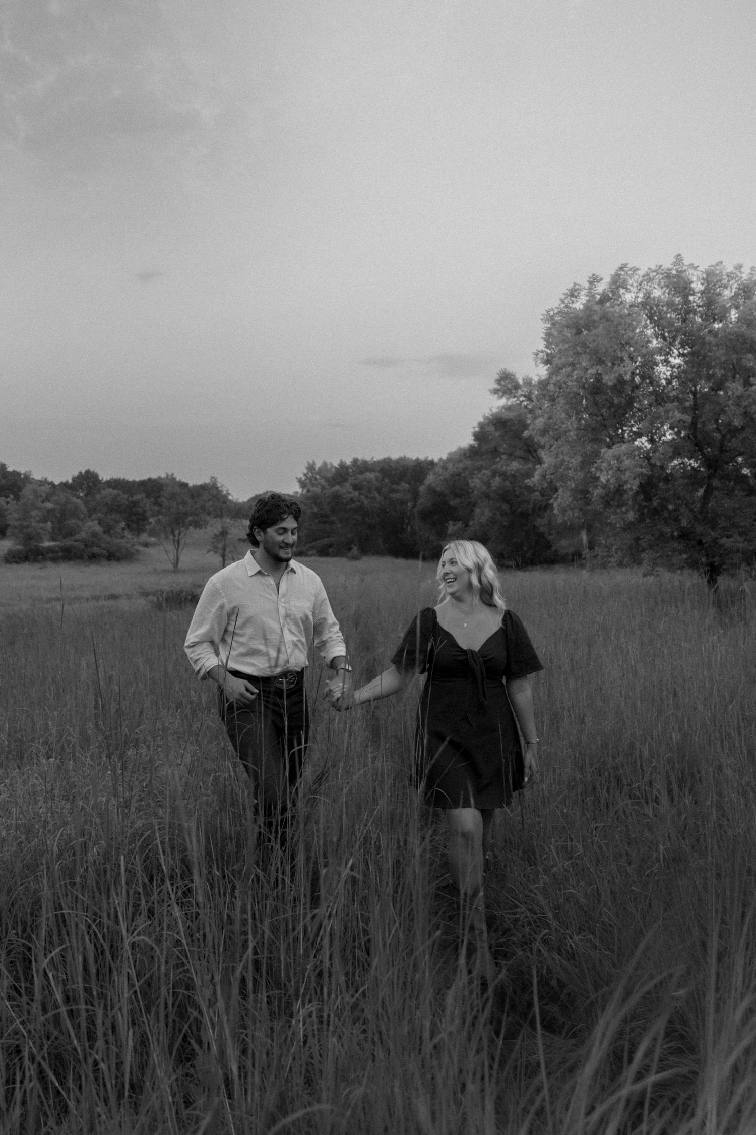 Couple holding hands walking through tall grass field in romantic black and white countryside photo.